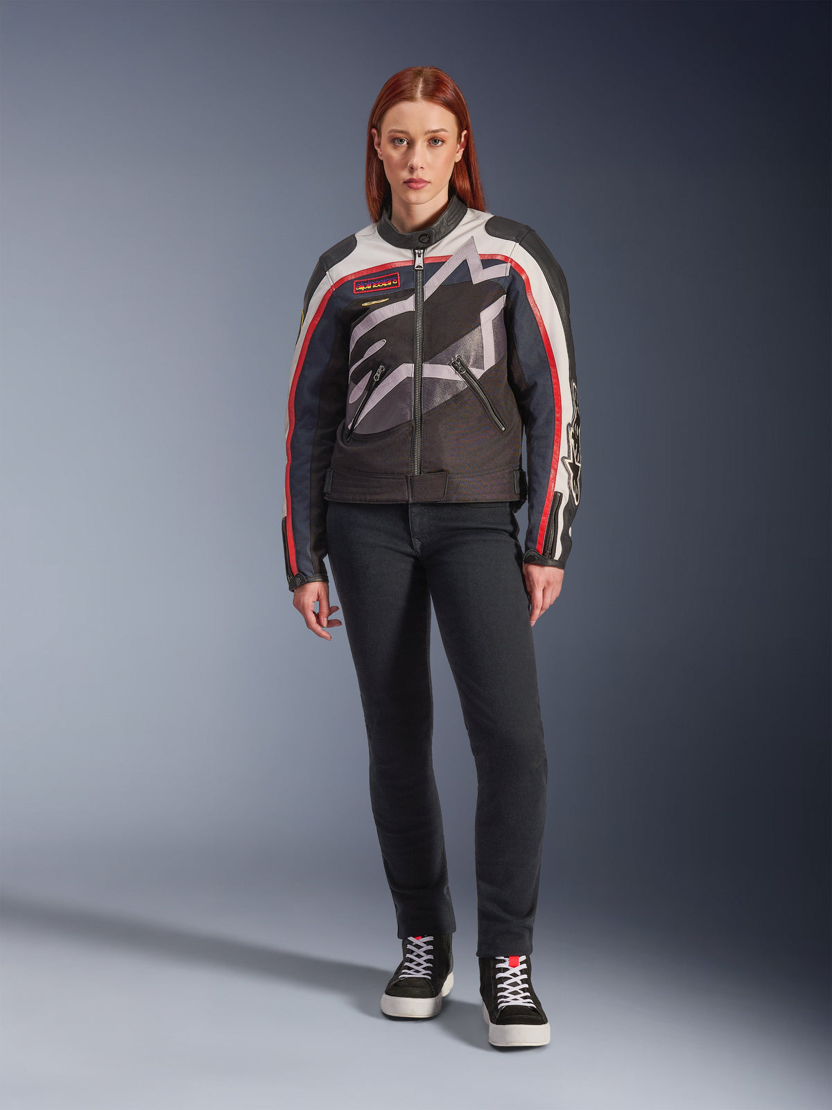 Vrouwelijk model draagt de Alpinestars Idles Jacket, motorjas voor dames, gebroken wit met vlamrood en zwart, voorzien van een groot rondomlopend sterlogo en vintage-geïnspireerde branding, gecombineerd met een donkere slim-fit motorbroek en zwarte high-top sneakers, urban eigentijdse motoruitrusting