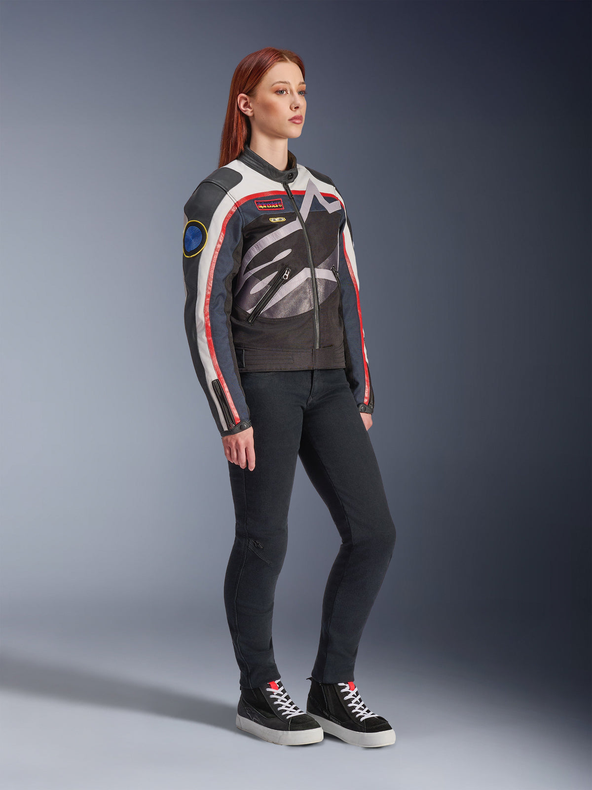 Alpinestars Idles Damesjas, motorjas, Off White, Flame Red en zwart, met een groot rondom lopend Alpinestars-logo en vintage racing-geïnspireerde patches, gedragen met een zwarte broek en hoge sneakers, eigentijdse urban motorkleding
