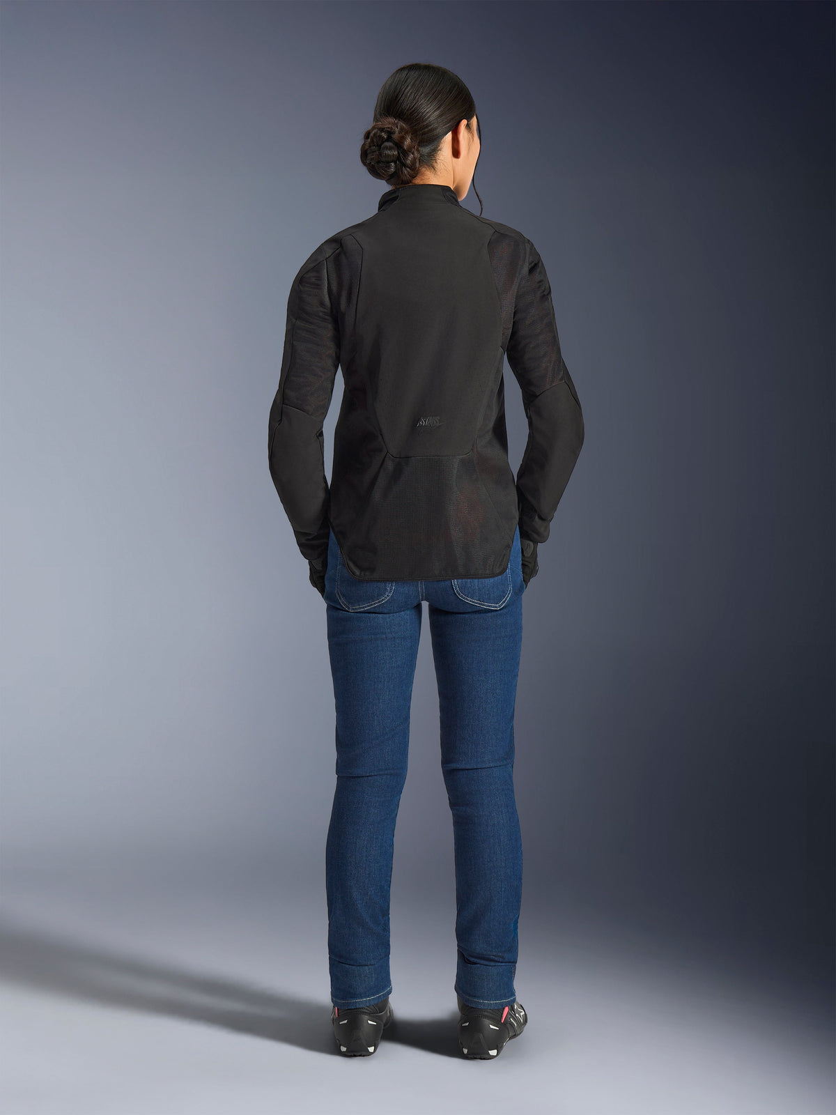 Alpinestars Stella Aeroshell Airflow damesjas, Black Anthracite, achteraanzicht op een model, voorzien van lichtgewicht softshell chassis met laser-geperforeerde meshpanelen, verlengd achterprofiel voor dekking, sportieve rijpasvorm, gecombineerd met een blauwe jeans