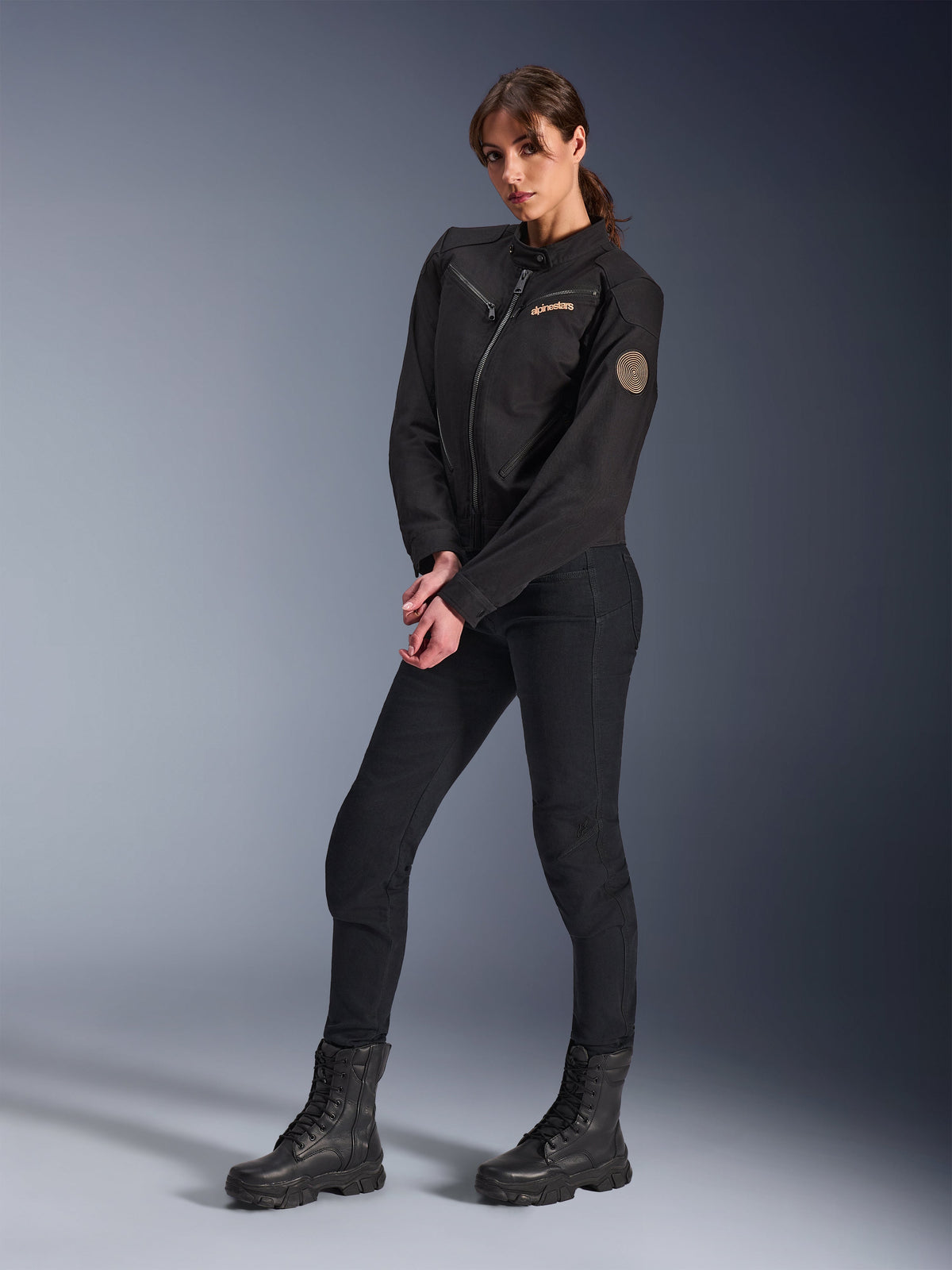 Female model wearing the Alpinestars Woman Stella Grace Jacket, Black Urban Motorcycle Jacket, styled with black riding pants and boots, workwear-inspired design featuring a bronze embroidered logo on the chest and a circular spiral patch on the sleeve