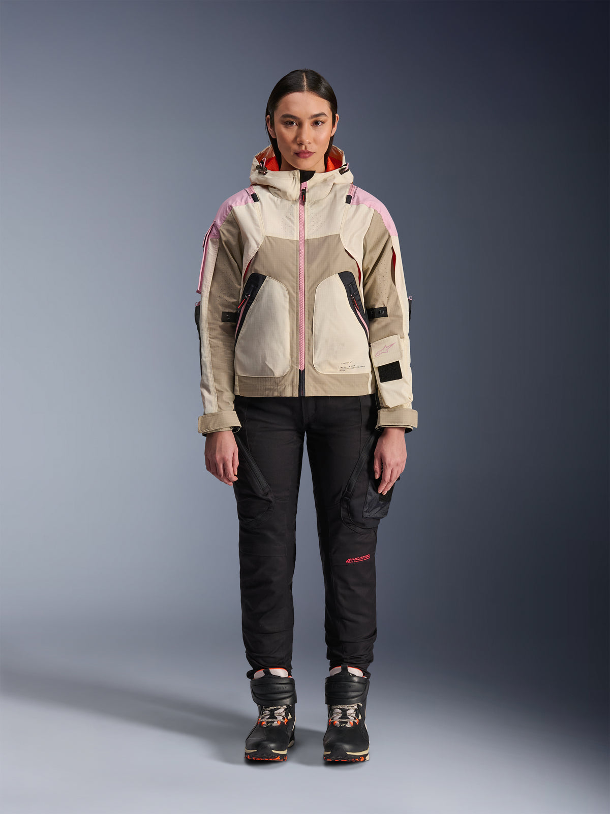 Vrouwelijk model draagt de Alpinestars Molly Air Jas in Sand Pink Almond, gecombineerd met een zwarte technische motorbroek en laarzen, vooraanzicht van het hele lichaam, urbane motorjas met utility-geïnspireerd ontwerp en roze accenten