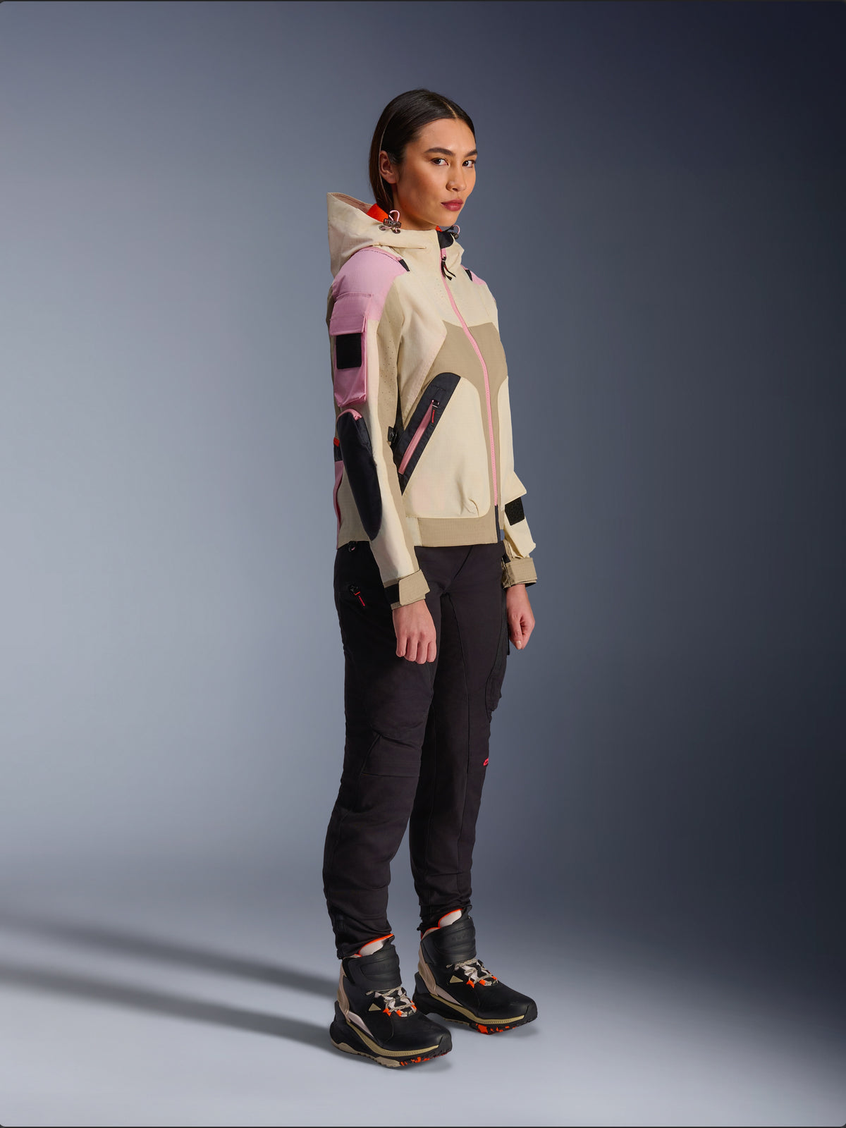 Vrouwelijk model draagt de Alpinestars Molly Air Jacket in Sand Pink Almond, gecombineerd met een donkere technische broek en hoge motorschoenen, urban motorjas voorzien van een capuchon en meerkleurige panelen, eigentijds op streetwear geïnspireerd ontwerp