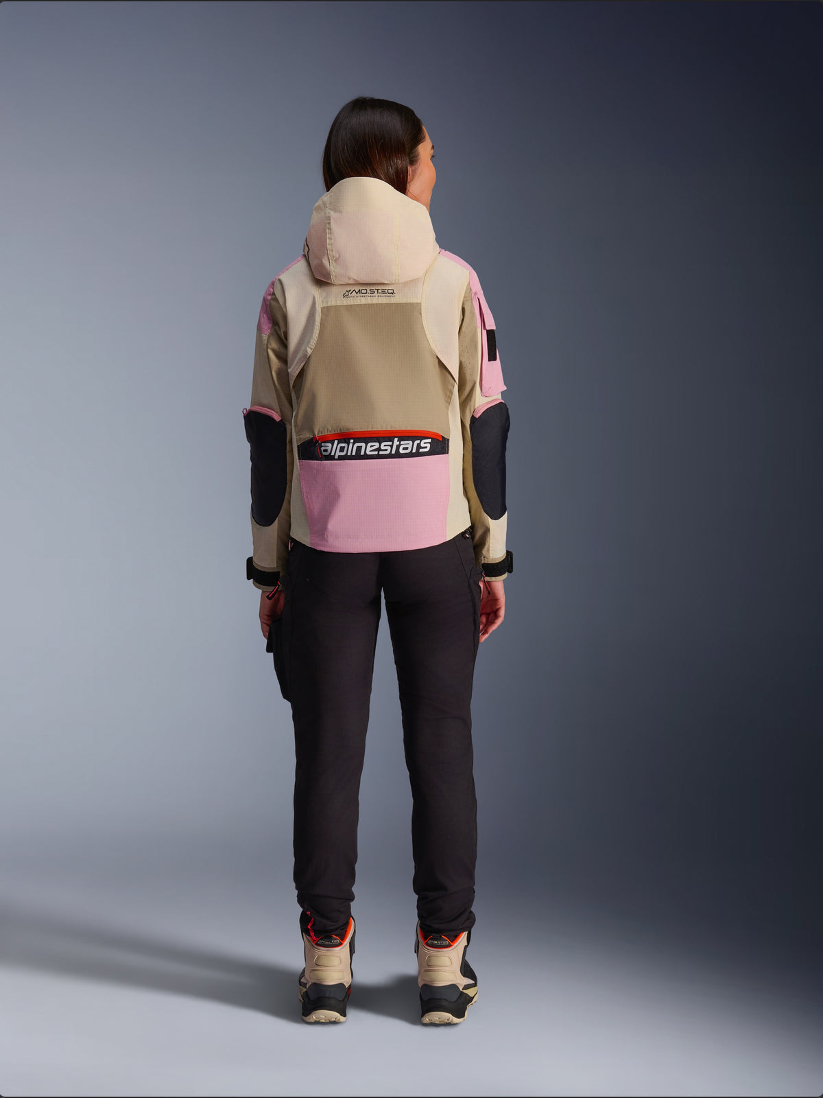 Vrouwelijke motorrijder van achteren gezien die de Alpinestars Molly Air Jacket draagt in Sand Pink Almond, met een zandkleurige capuchon en roze panelen met zwarte accenten, gecombineerd met een zwarte motorbroek en zandkleurige motorlaarzen, urban hedendaagse motorkleding