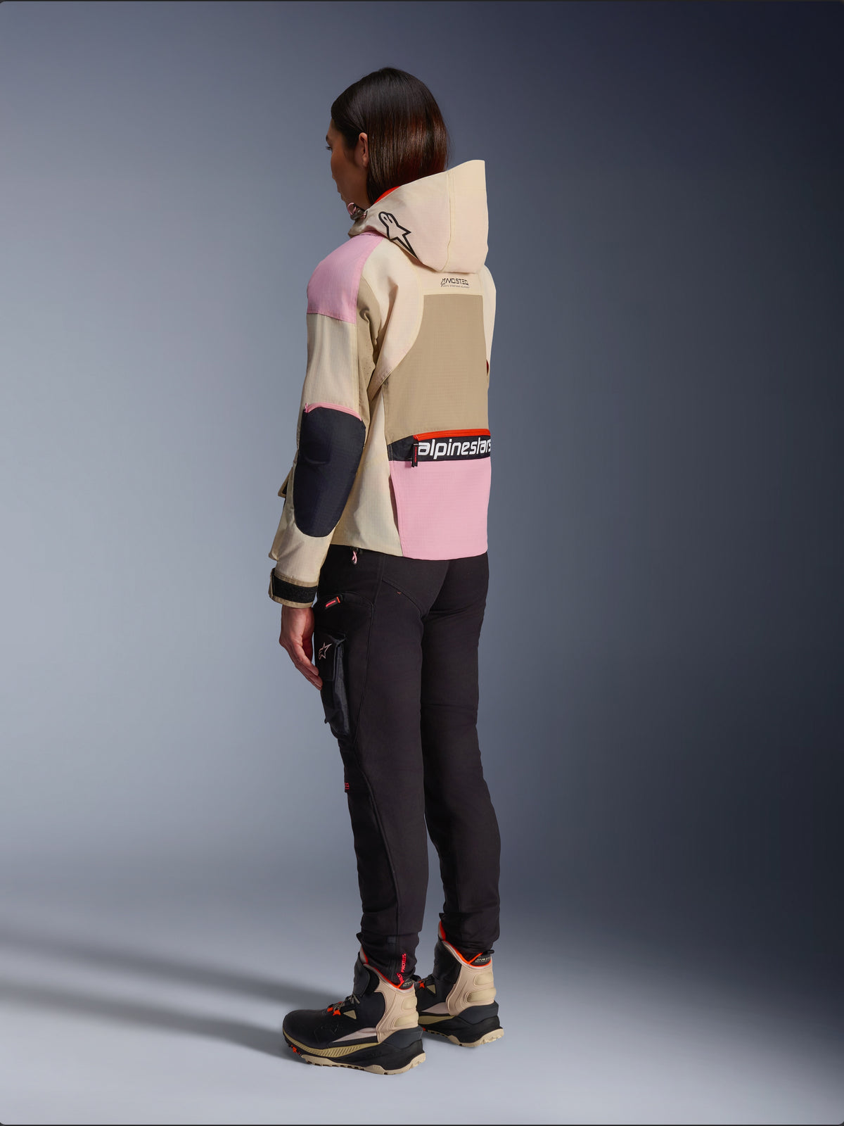 Vrouwelijke motorrijder die de Alpinestars Molly Air Jacket draagt in Sand Pink Almond, met een meerkleurig beige en roze ontwerp, achteraanzicht van opzij met zwarte motorbroek en bijpassende technische laarzen, urban motorstijl met capuchon.
