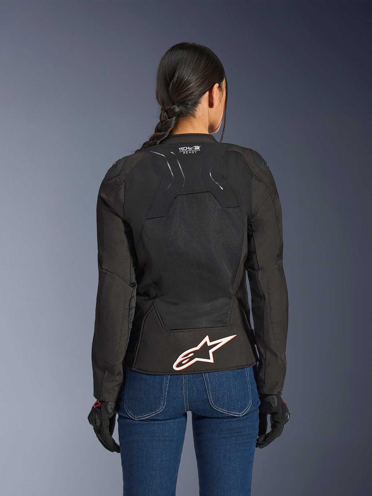 Alpinestars Stella SMX Air jas, achteraanzicht op vrouwelijk model, zwart textiel met high-flow mesh panelen, 'TECH-AIR READY' branding op de bovenrug, groot wit Alpinestars-logo met rode omtrek op de onderrug, gecombineerd met een blauwe spijkerbroek en zwarte motorhandschoenen