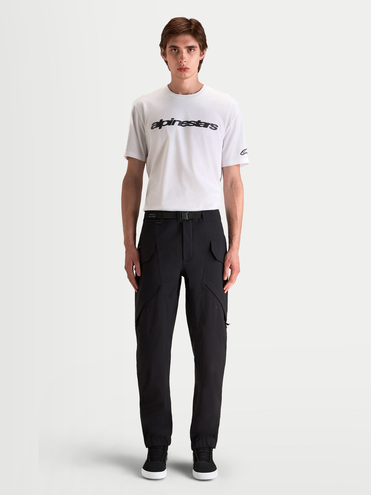 Alpinestars Juggernaut V2 Drystar broek, waterdichte urban motorbroek voor heren in zwart, vooraanzicht op model met witte t-shirt en casual motorlaarzen, voorzien van cargozakken en een verstelbare riem