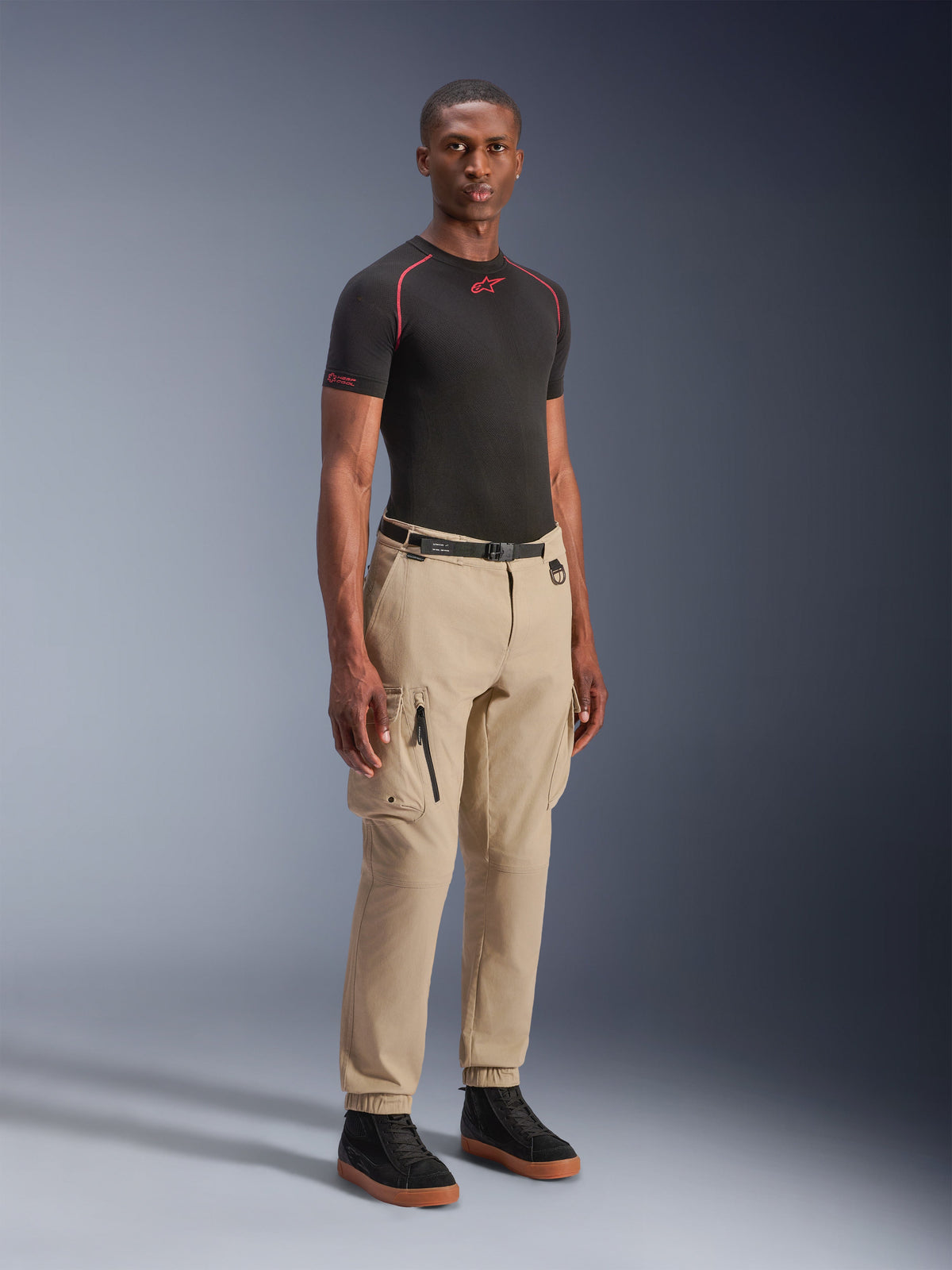 Model draagt Alpinestars Flex-AST Cargo Pants in Light Beige, regular tapered fit met elastische zomen, grote cargozakken op de dijen, zwarte riem, gecombineerd met een zwarte basislaag met korte mouwen en zwarte sneakers, urban motorstijl