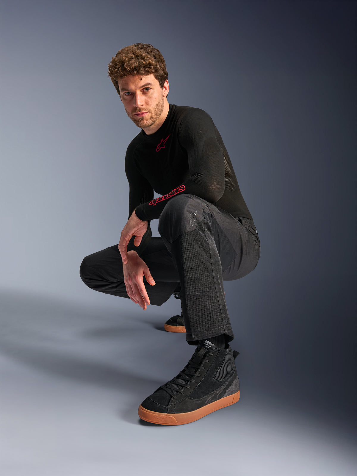 Alpinestars C-1 Air broek, urban motorbroek, zwart, model hurkend in een studio-setting, gedragen met een zwarte top met lange mouwen en zwarte sneakers met bruine zolen, voorzien van ademende mesh-panelen en een eigentijdse urban stijl