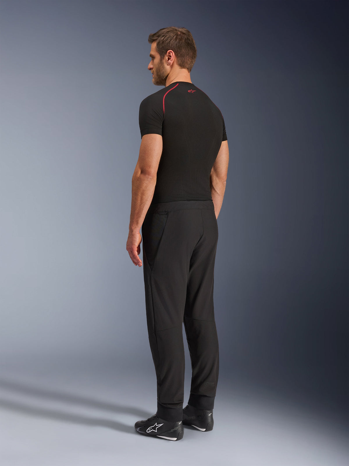 Alpinestars Aeroshell Airflow broek, zwarte motorbroek, lichtgewicht softshell textiel, gedragen met een zwart technisch shirt met korte mouwen en zwarte motorlaarzen, achteraanzicht in profiel toont de slanke pasvorm en ergonomische constructie.