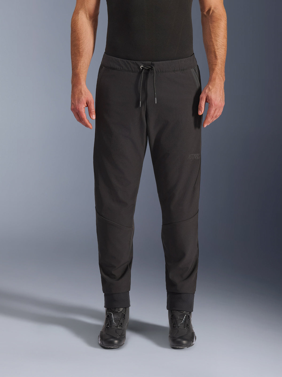 Alpinestars Aeroshell Airflow broek, zwarte motorbroek, vooraanzicht gedragen met zwarte motorlaarzen, voorzien van een taille met trekkoord en taps toelopende enkelmanchetten, lichtgewicht softshell-ontwerp met subtiele ton-sur-ton branding.