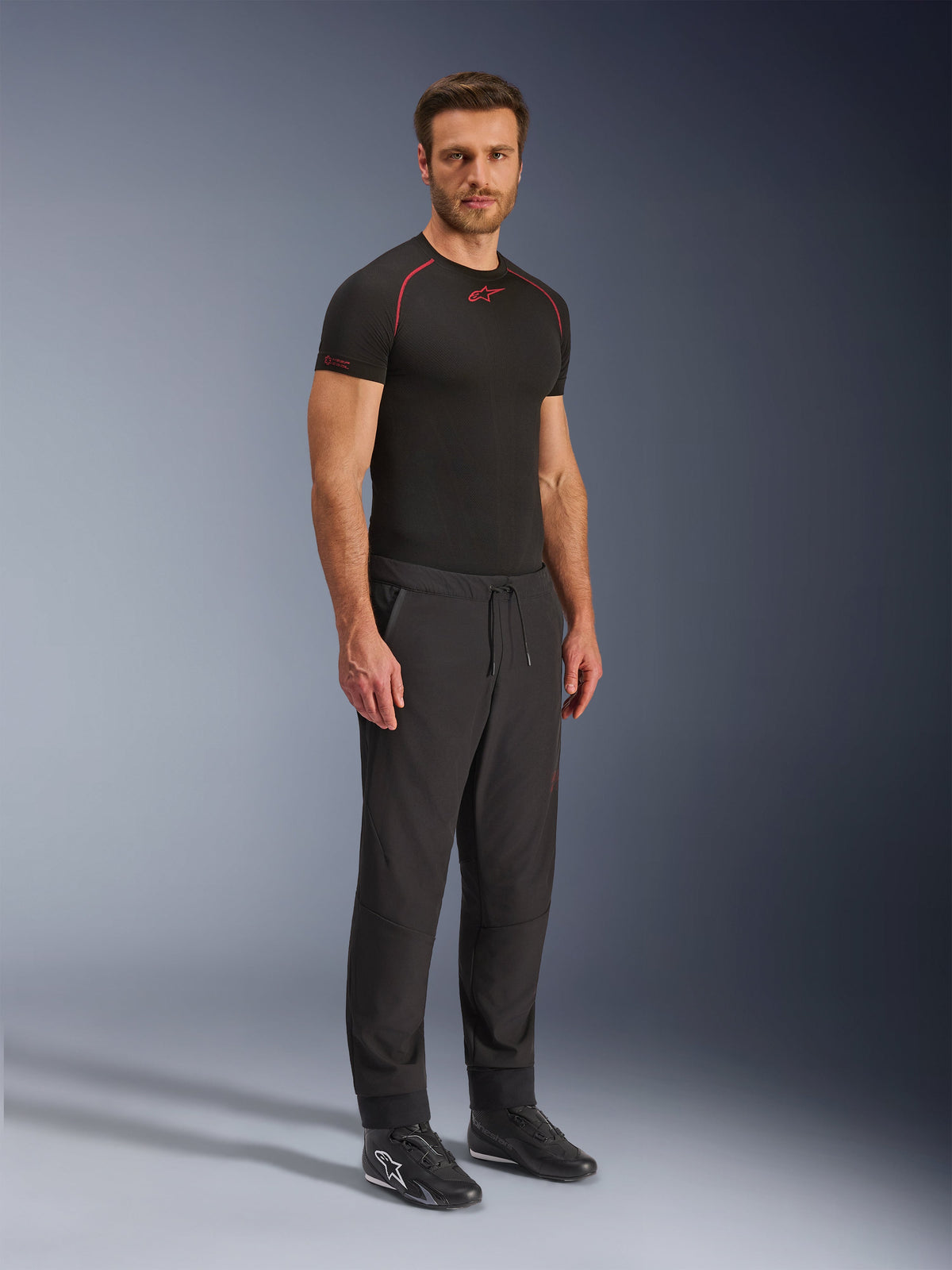 Alpinestars Aeroshell Airflow Broek, zwarte motorbroek, volledige studiofoto van een model die een softshell broek draagt met een trekkoord in de taille en elastische boorden, gecombineerd met een zwart Alpinestars base layer shirt en zwarte motorschoenen.
