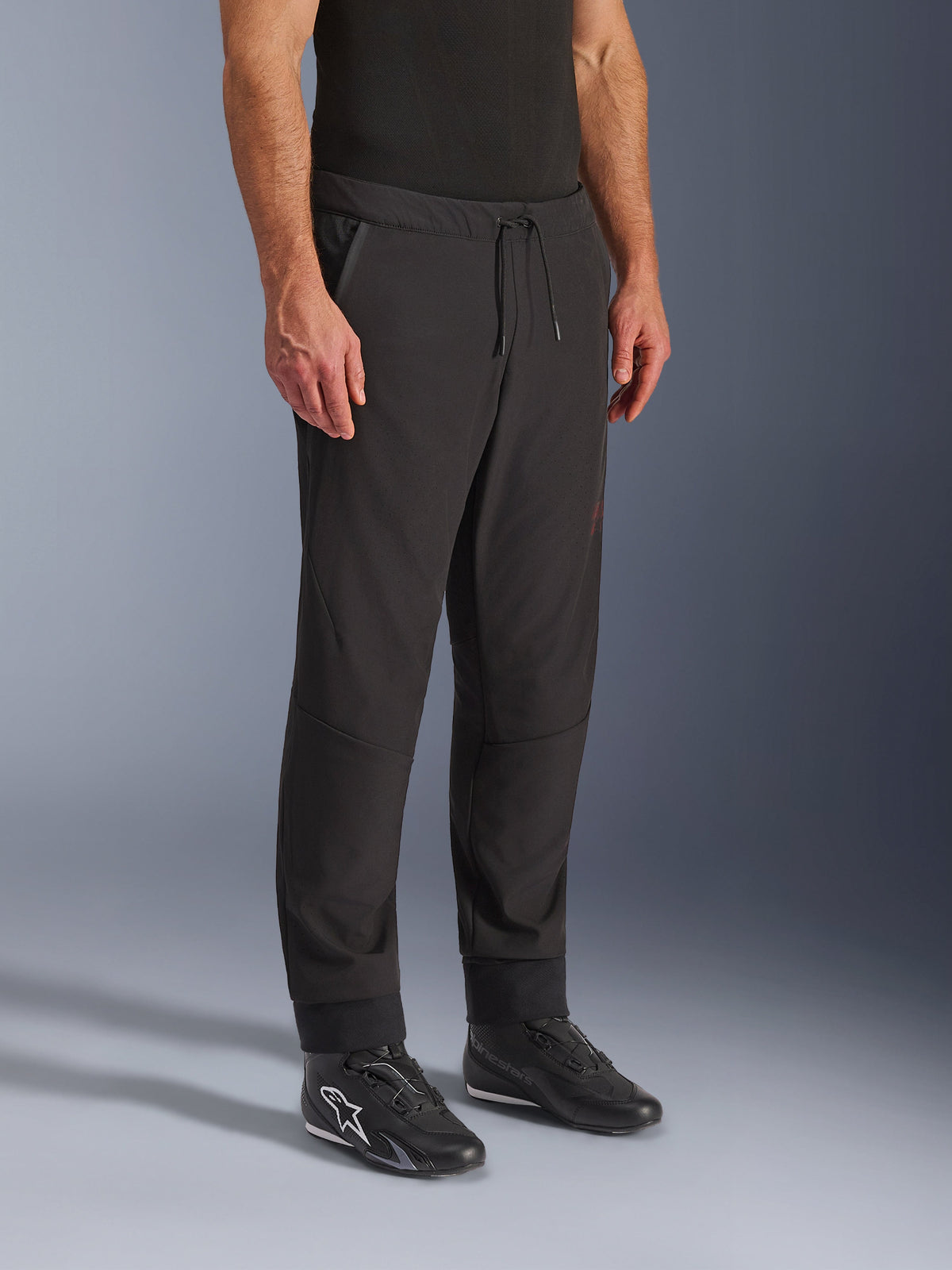 Alpinestars Aeroshell Airflow broek, zwarte motorbroek, gedragen door model met zwarte rijschoenen, voorzien van een tailleband met trekkoord, nauwsluitende enkelboorden en laser-geperforeerde softshell-stof voor maximale ventilatie