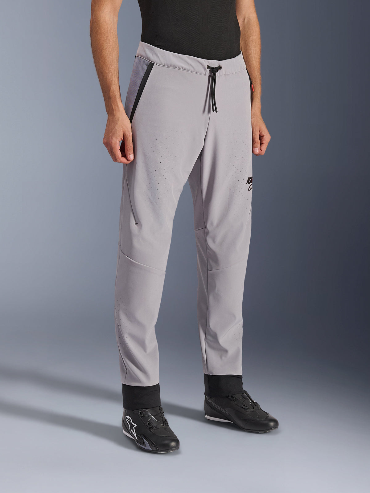 Alpinestars Aeroshell Airflow Broek, Mid Gray, lichtgewicht motorbroek met laser-geperforeerde ventilatiezones op de dijen en schenen, voorzien van een elastische tailleband met trekkoord en een slanke sportieve pasvorm, gedragen door een model met zwarte motorschoenen.
