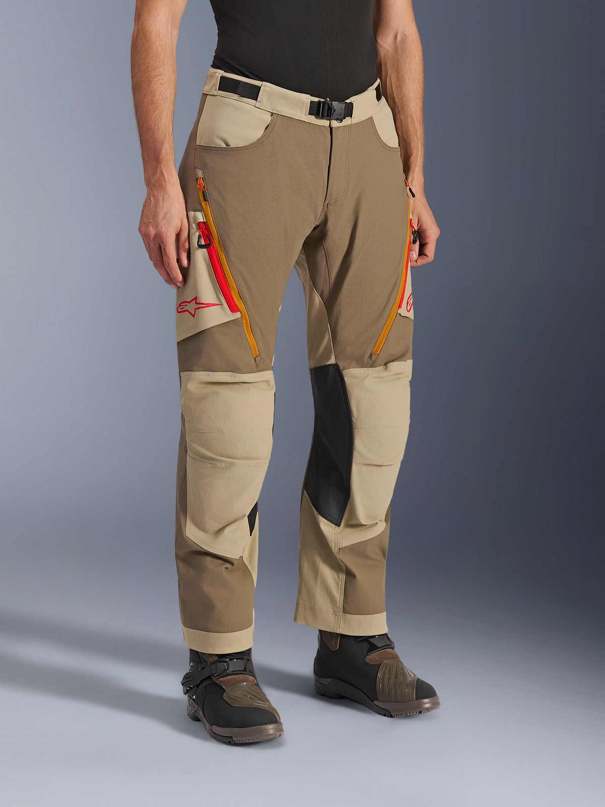 Alpinestars Maxdura Dual Pants, Walnut Aluminum bruine en khaki adventure motorbroek, met oranje en rode ventilatieritsen op de dijen, verstevigde zwarte leren binnenkniepanelen en rood Astars-logo, gedragen met technische laarzen.