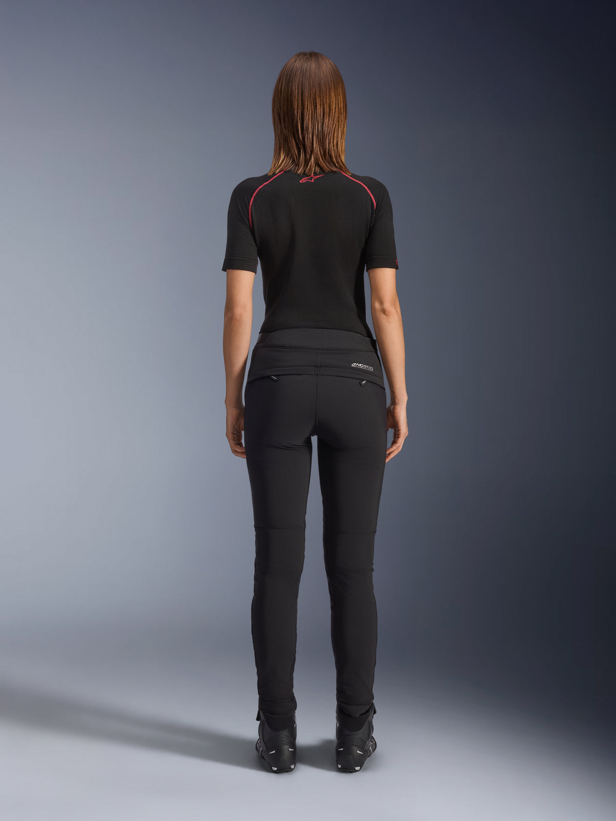 Alpinestars Alamosa damesbroek, zwart, achteraanzicht met een slanke textielconstructie, achterzakken met ritssluiting en reflecterende MO.ST.EQ.-branding op de achterpas, gedragen met een zwarte basislaag met korte mouwen en zwarte motorlaarzen