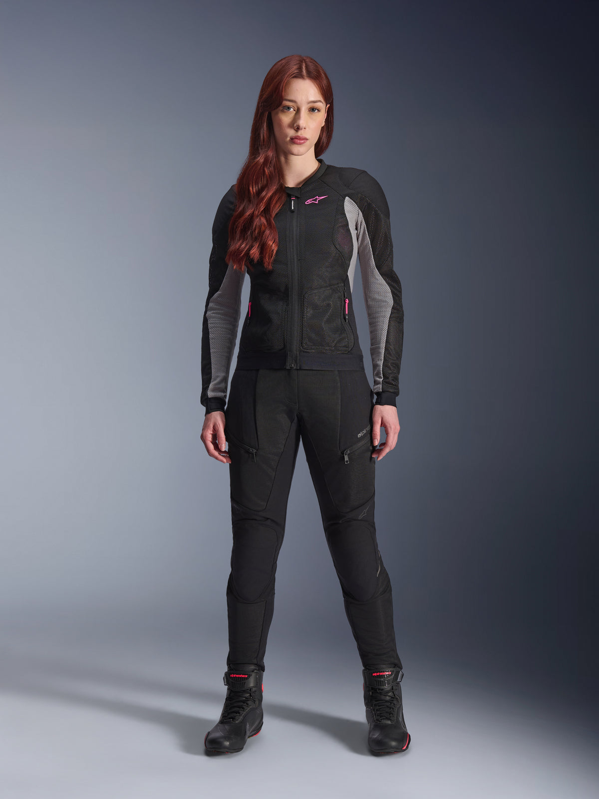 Alpinestars Stella Troop-Air Pants, dames motorbroek in het zwart, voorzien van meshpanelen en zakken met rits, gecombineerd met een bijpassende zwart-grijze mesh jas met roze accenten en zwarte motorlaarzen, complete stedelijke motoroutfit.
