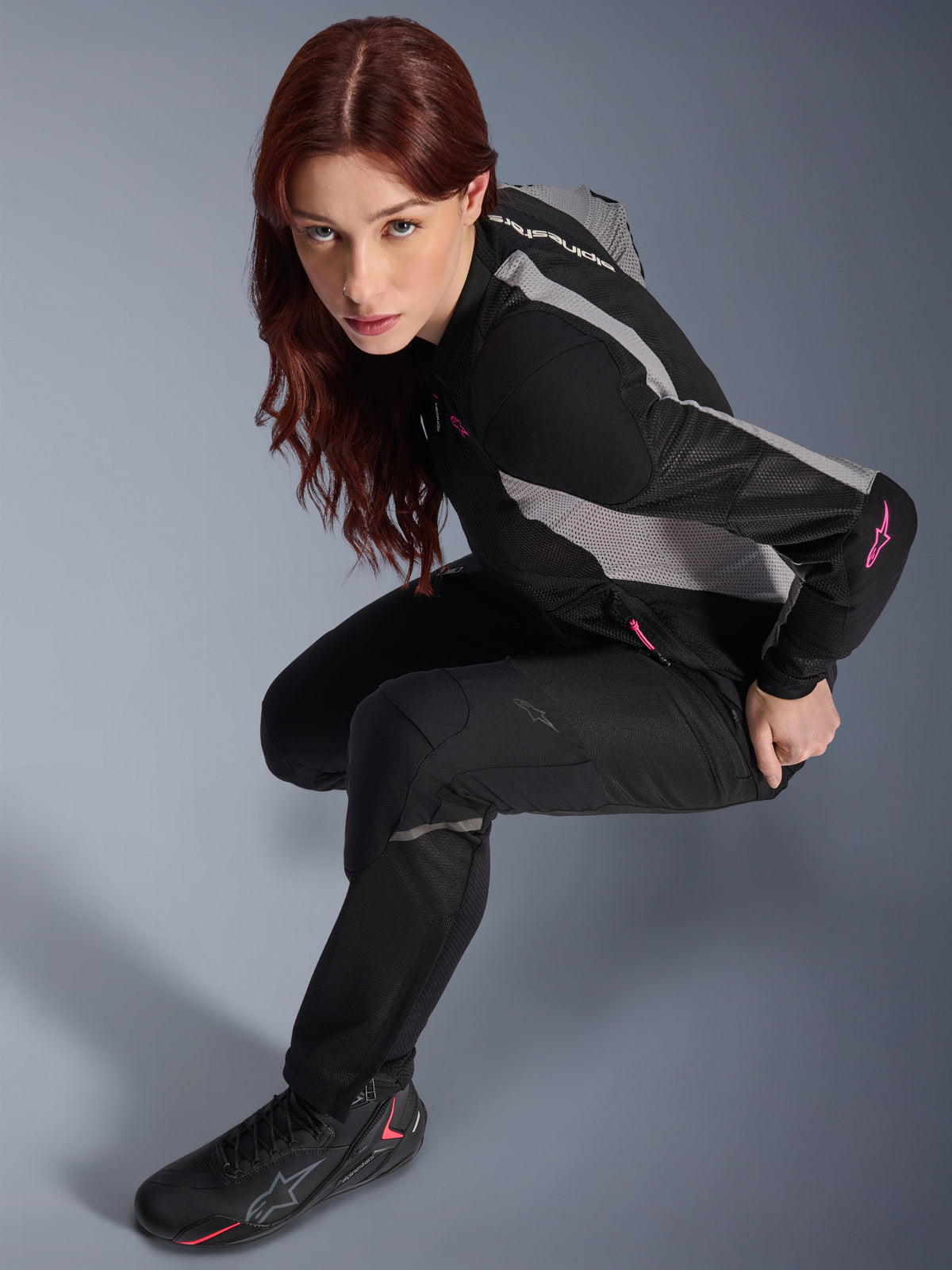 Vrouw die de Alpinestars Stella Troop-Air broek draagt, zwarte lichtgewicht geventileerde textielbroek, gecombineerd met een zwart en grijs mesh jack met roze accenten en zwarte motorlaarzen, studioportret, urban motoruitrusting