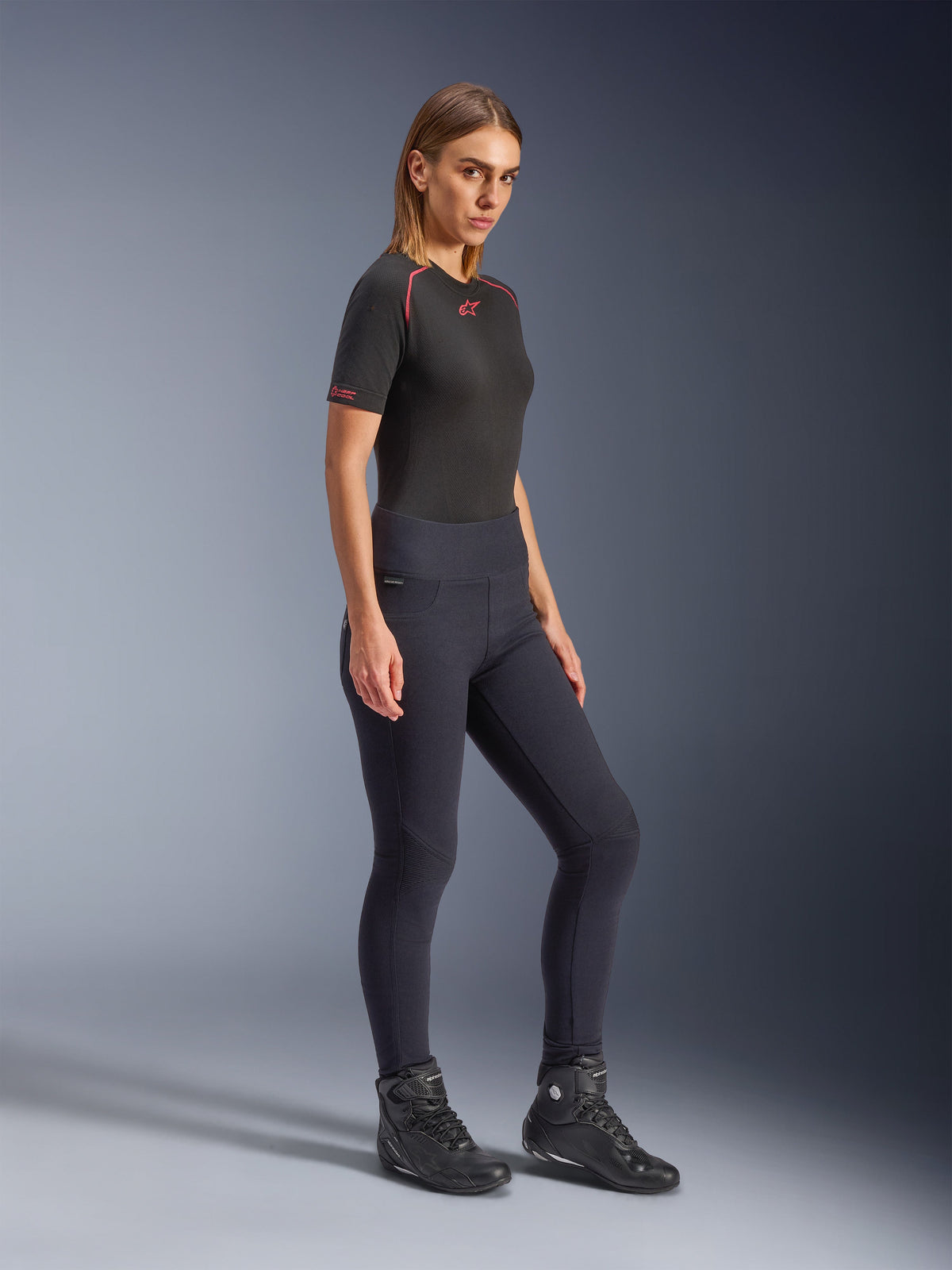Model draagt Alpinestars Stella Flex-AST dameslegging in het zwart, skinny pasvorm met hoge taille en harmonicavormige kniepanelen, gecombineerd met een zwart Alpinestars-shirt met korte mouwen en zwarte motorlaarzen, volledige studio-opname.