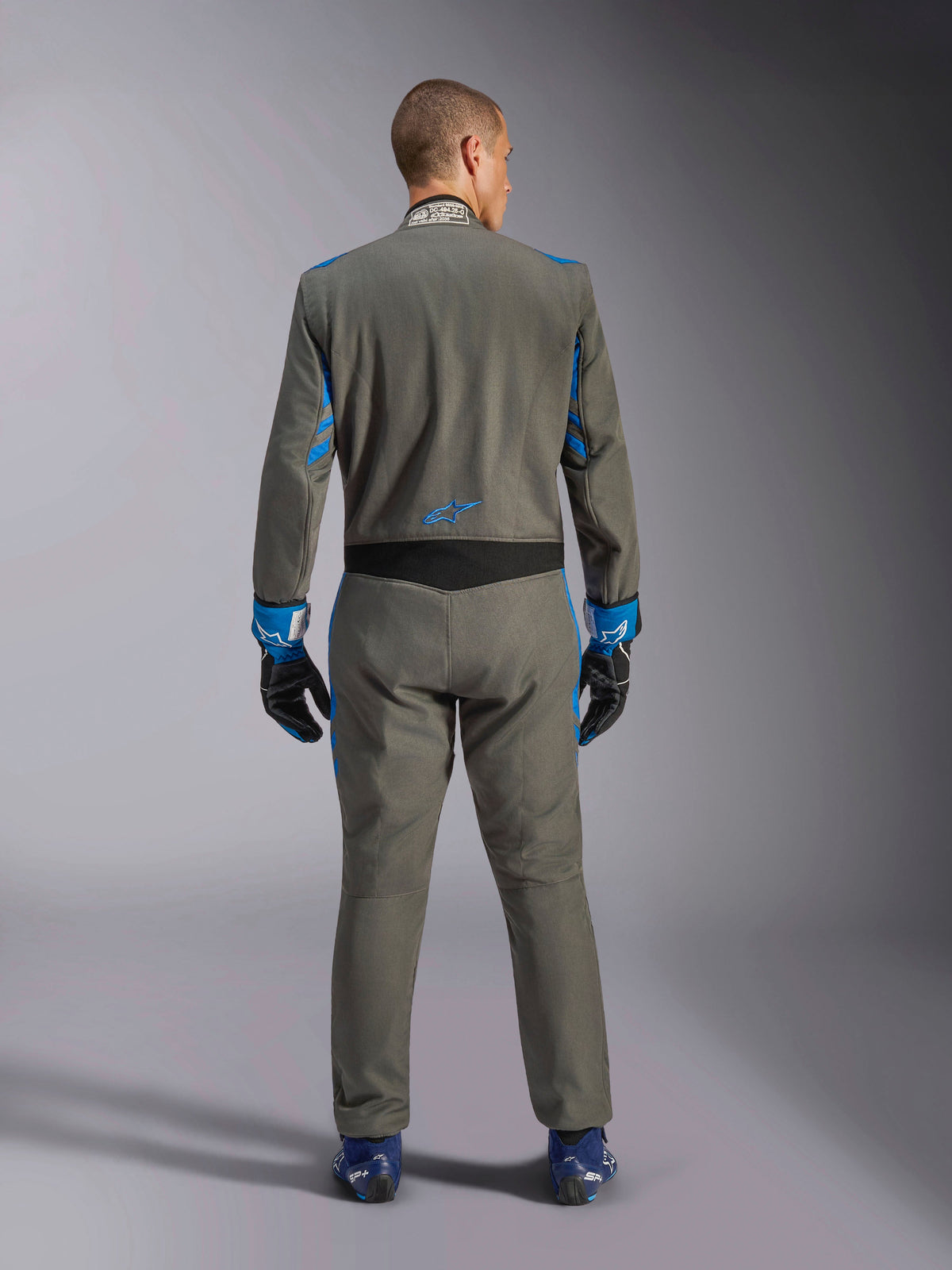 Alpinestars GP Race V3 Suit FIA, Back View, Anthracite Blue with vibrant blue details on the arms and shoulders, featuring blue star logo on lower back, worn with matching racing gloves and blue shoes, professional auto racing suit