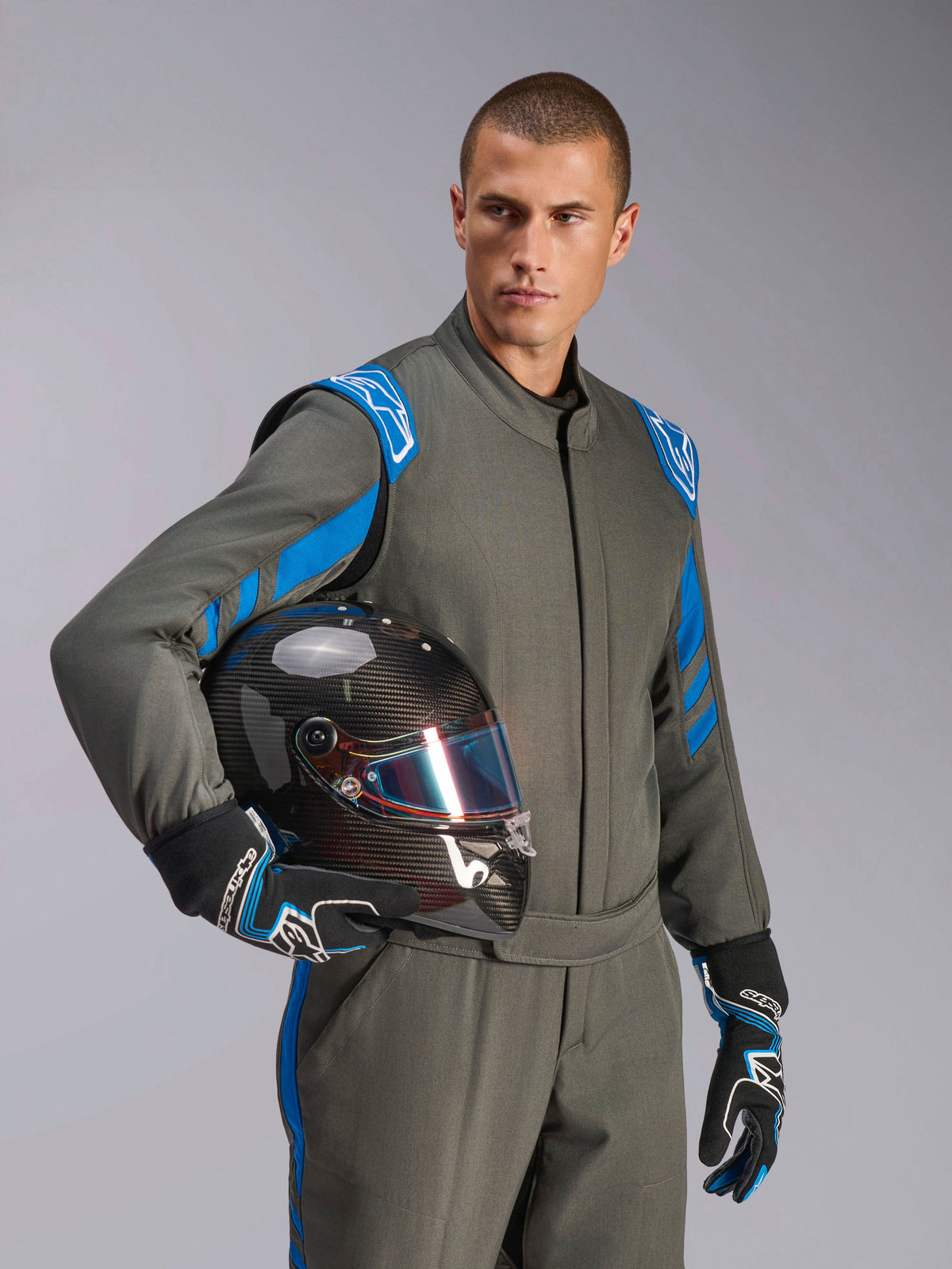 Male driver wearing Alpinestars GP Race V3 Suit FIA in Anthracite Blue, holding a carbon fiber racing helmet with an iridescent visor, professional auto racing gear with blue shoulder accents and matching racing gloves.