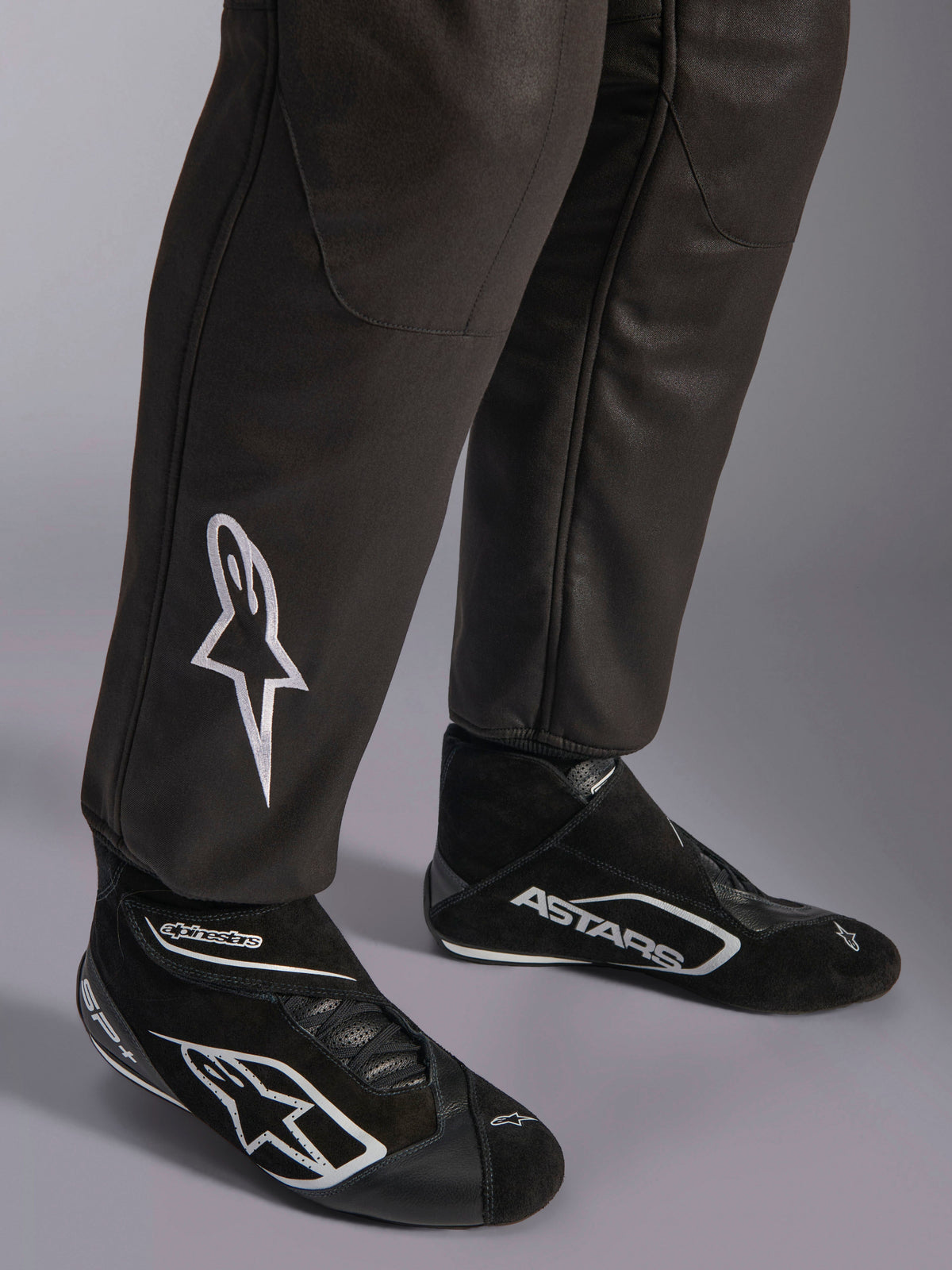 Alpinestars GP Race V3 Suit FIA, close-up of lower legs and racing boots, Black with Silver accents, featuring white embroidered star logo on the shin and ribbed ankle cuffs, professional auto racing protective gear.