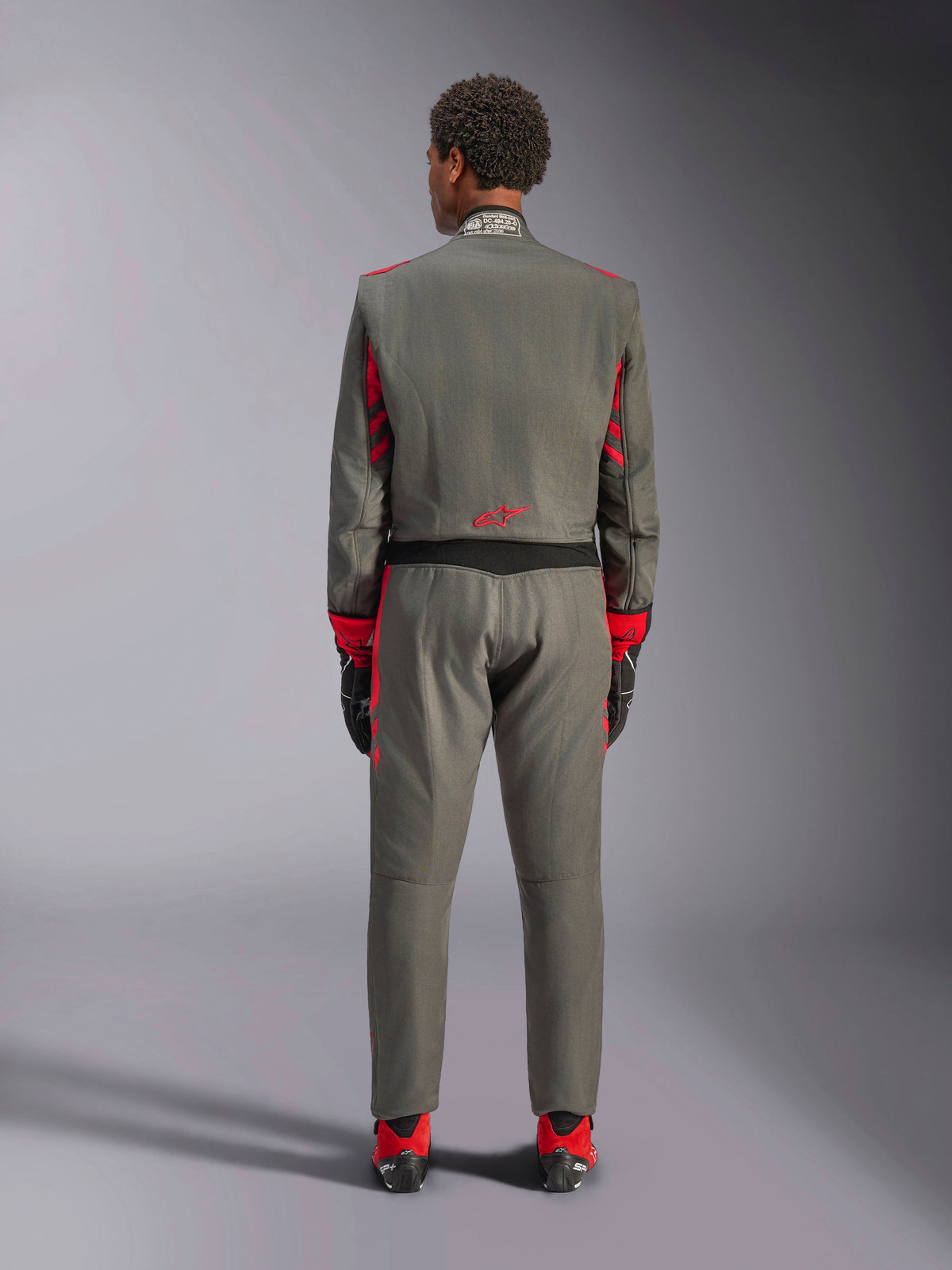 Rear view of a model wearing the Alpinestars GP Race V3 Suit in Anthracite Red, professional auto racing suit with red accents on the sleeves and legs, paired with red racing boots and black gloves, technical motorsport apparel shown in a studio setting.