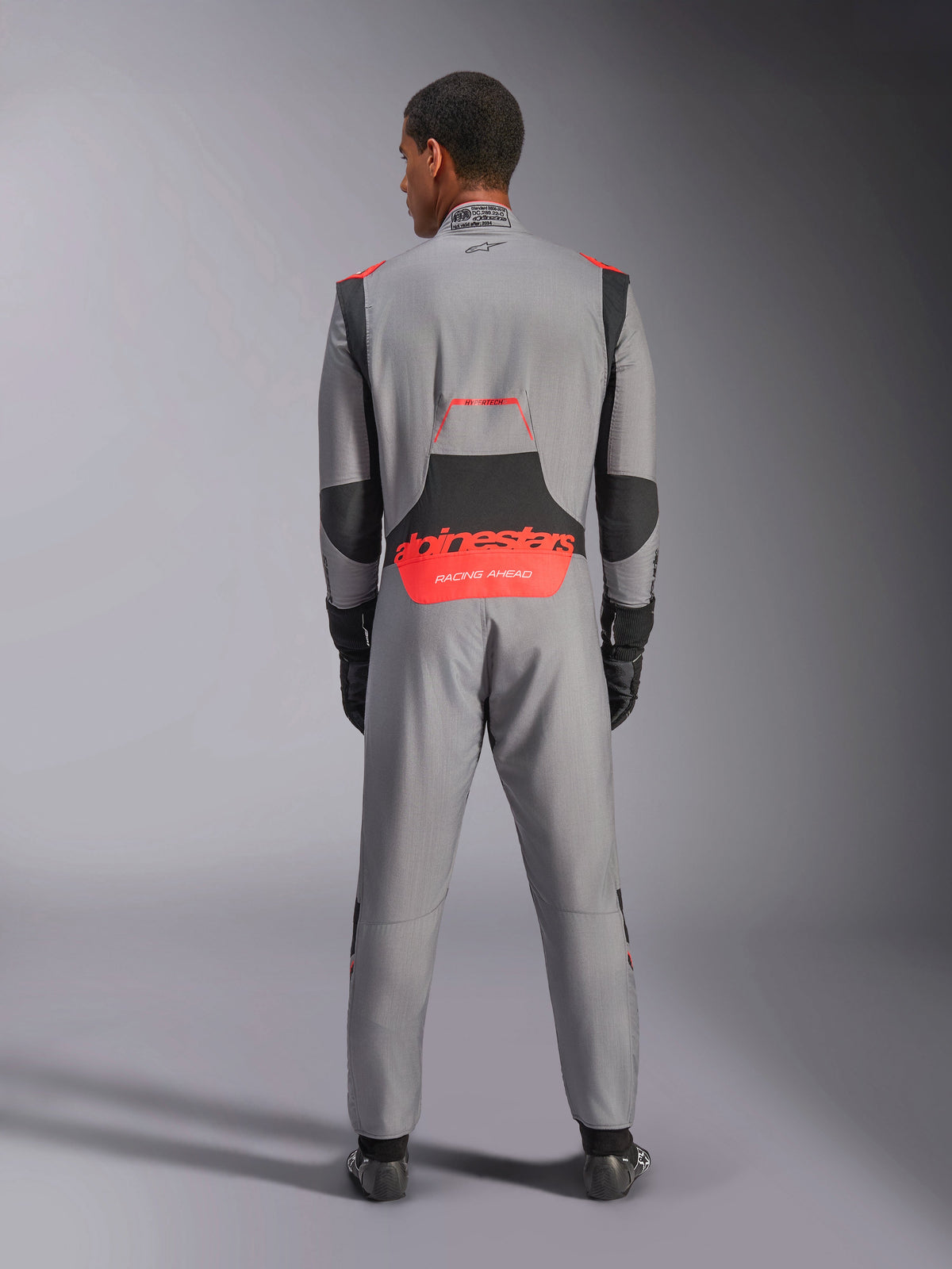 Alpinestars Hypertech V3 Suit, professioneel autoracepak, middengrijs met zwarte en fluo-rode accenten, achteraanzicht op een model met rode Alpinestars-branding op de onderrug, lichtgewicht Nomex-constructie voor circuitprestaties.
