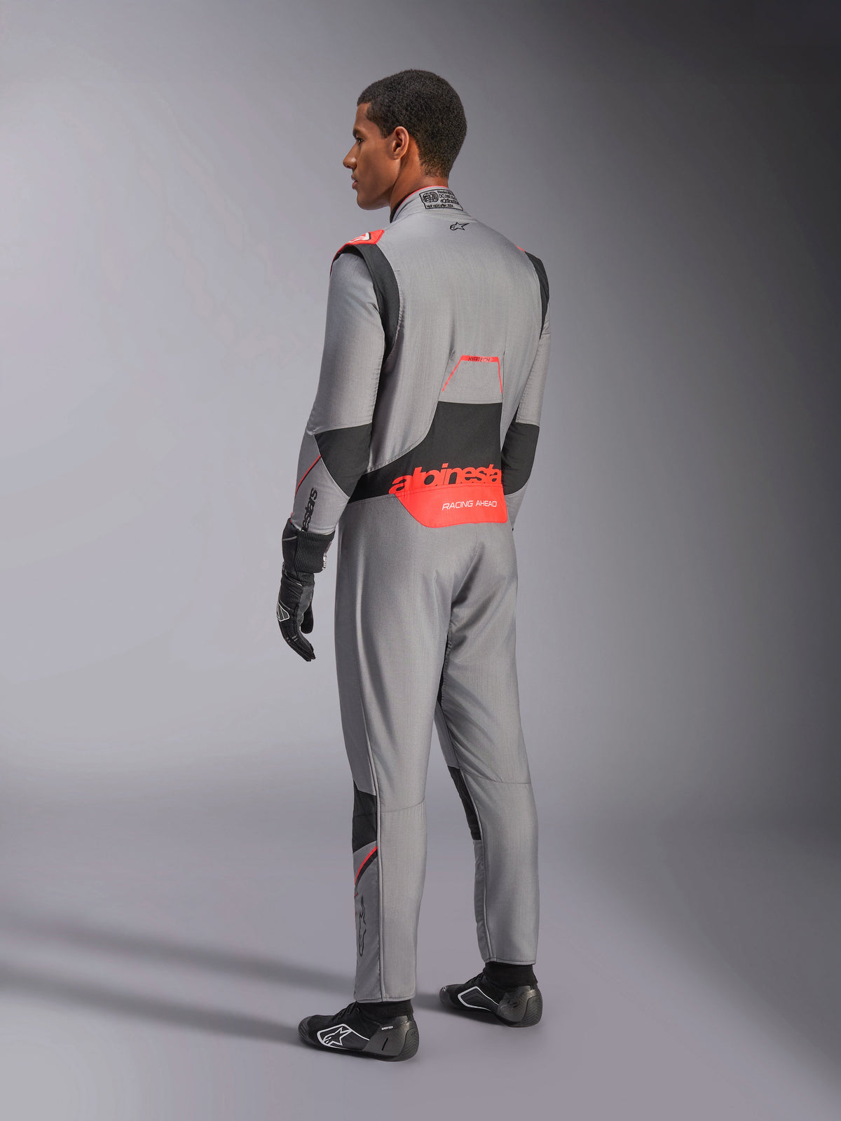 Alpinestars Hypertech V3 Suit in Mid Gray Black Red Fluo, driekwart achteraanzicht, met opvallende rode Alpinestars-branding op de onderrug, zwarte stretchpanelen en een slanke atletische pasvorm voor professionele autoraces.
