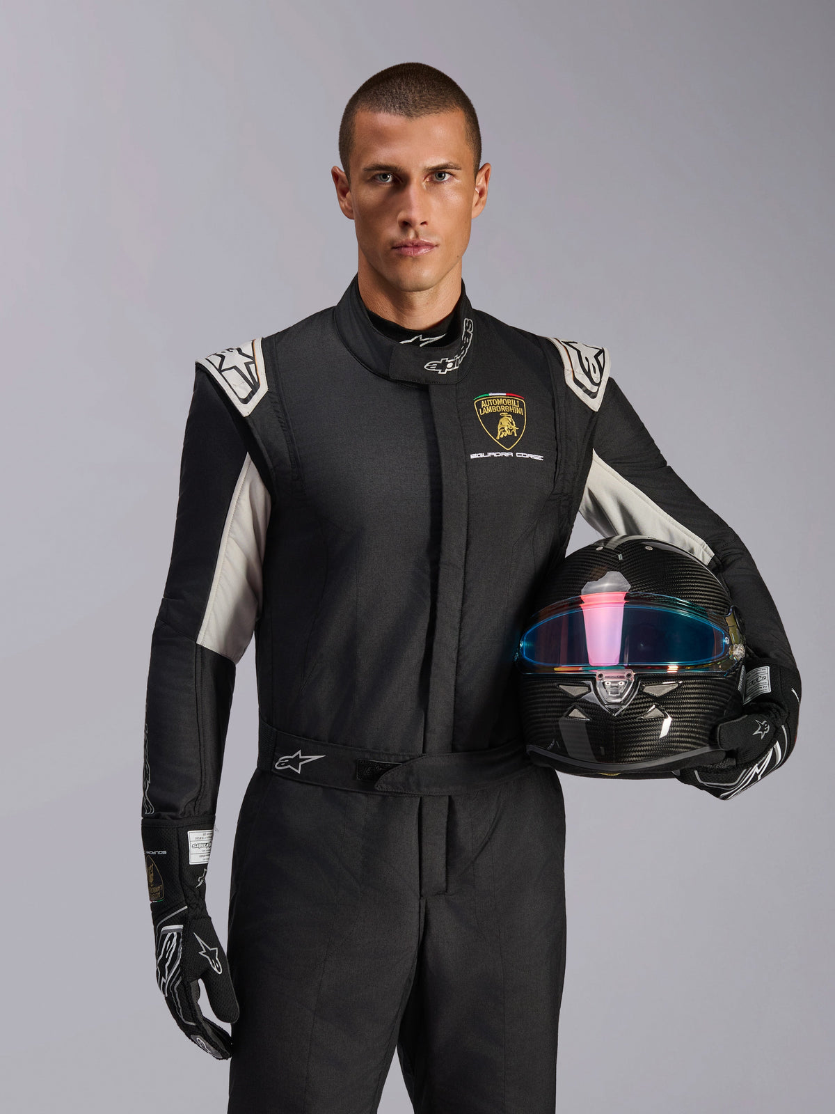 Male model wearing Limited Edition Lamborghini GP Tech V4 Suit, black racing suit with silver accents and Lamborghini Squadra Corse logo, holding a carbon fiber helmet, black racing gloves with white logos, professional auto racing gear