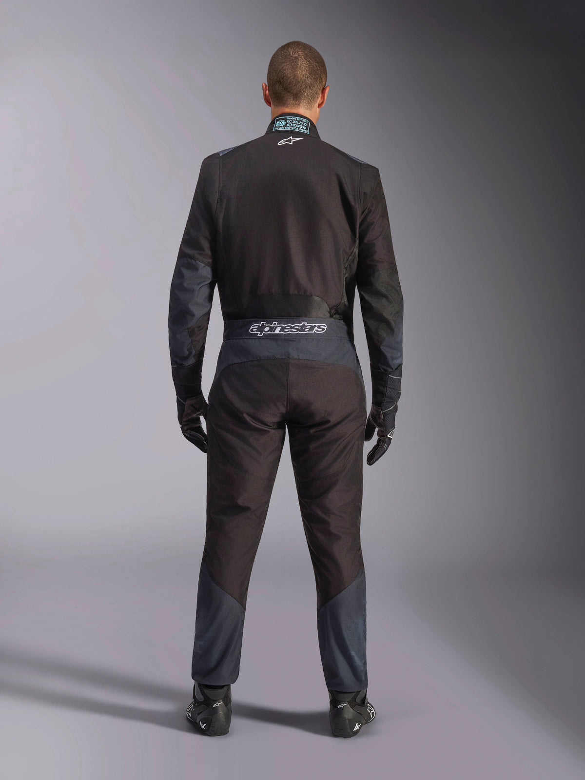 Alpinestars KMX-5 V3-pak, kartpak, zwart en Dark Shadow, achteraanzicht van een coureur die het volledige racepak draagt met bijpassende handschoenen en schoenen, voorzien van Alpinestars-branding op de tailleband en ergonomische panelen.
