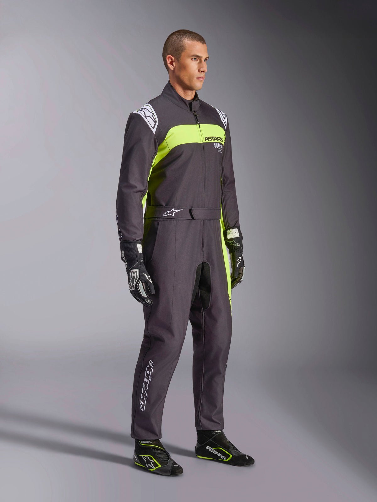 Male model wearing Alpinestars KMX-9 V3 Graphic 3 Suit in Black and Yellow Fluo, professional karting gear with vibrant neon yellow panels and white logo accents, accessorized with coordinated racing gloves and shoes, set against a studio background.