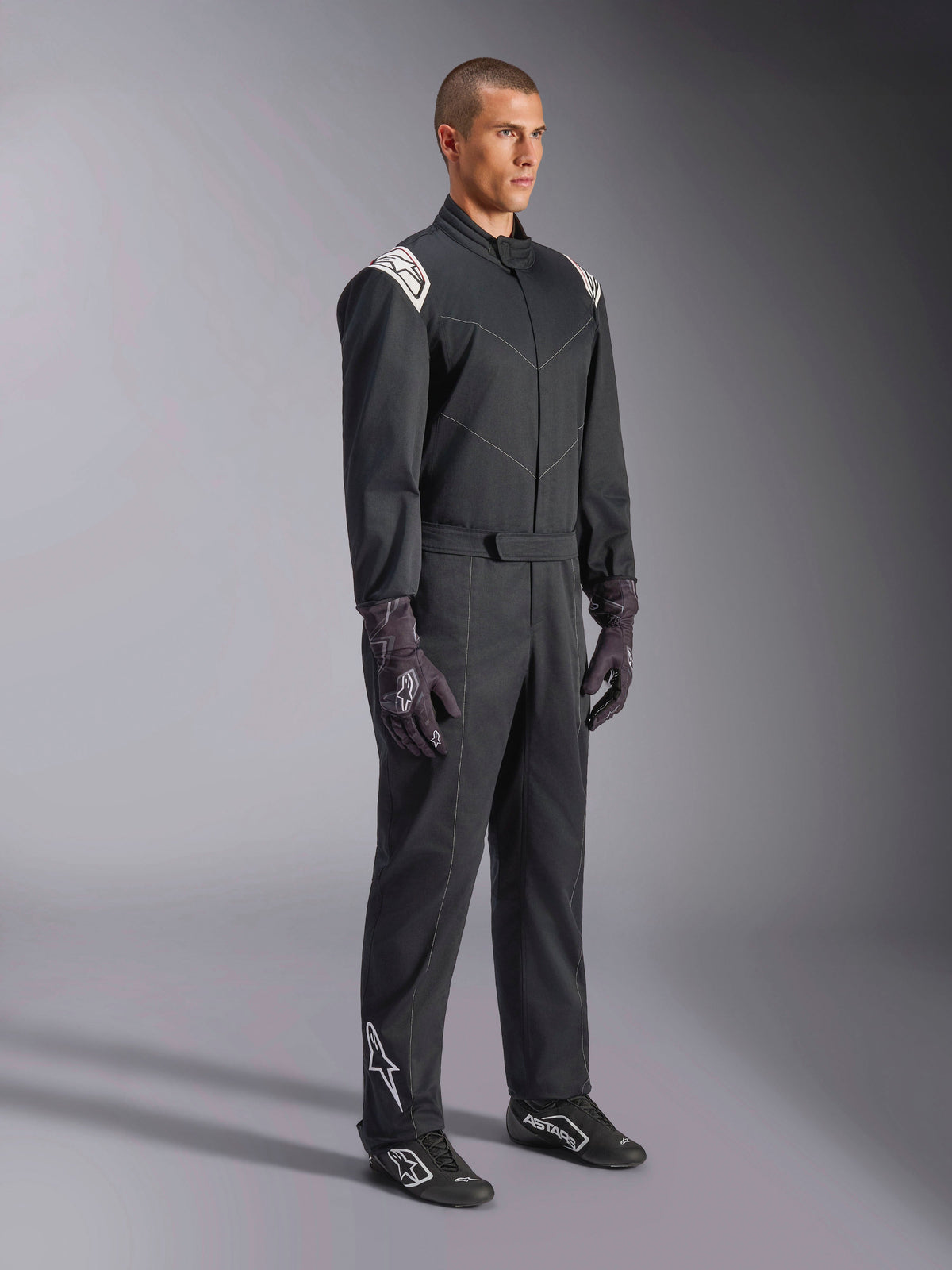 Alpinestars Kart Indoor V2 Suit, zwart eendelig kartpak, gedragen door een model met racehandschoenen en schoenen, voorzien van witte schouderaccenten en een wit Astars-logo op het onderbeen, professionele motorsportkleding voor indoor karten
