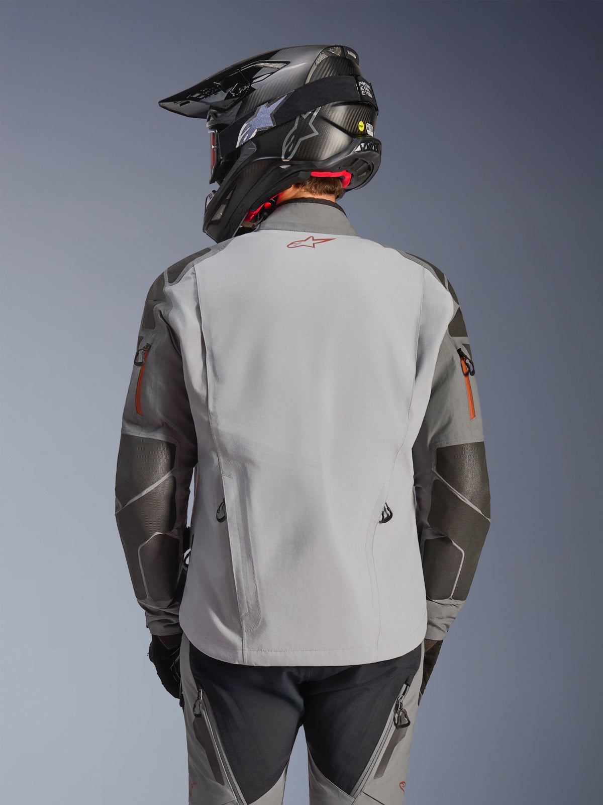 Alpinestars Nazca 3L GORE-TEX PRO Jas, adventure motorjas, achteraanzicht, donkergrijs en middengrijs, voorzien van Superfabric-verstevigingen op de ellebogen en schouders, rood logo op de rug, gedragen met bijpassende adventure-uitrusting en een helm van koolstofvezel.