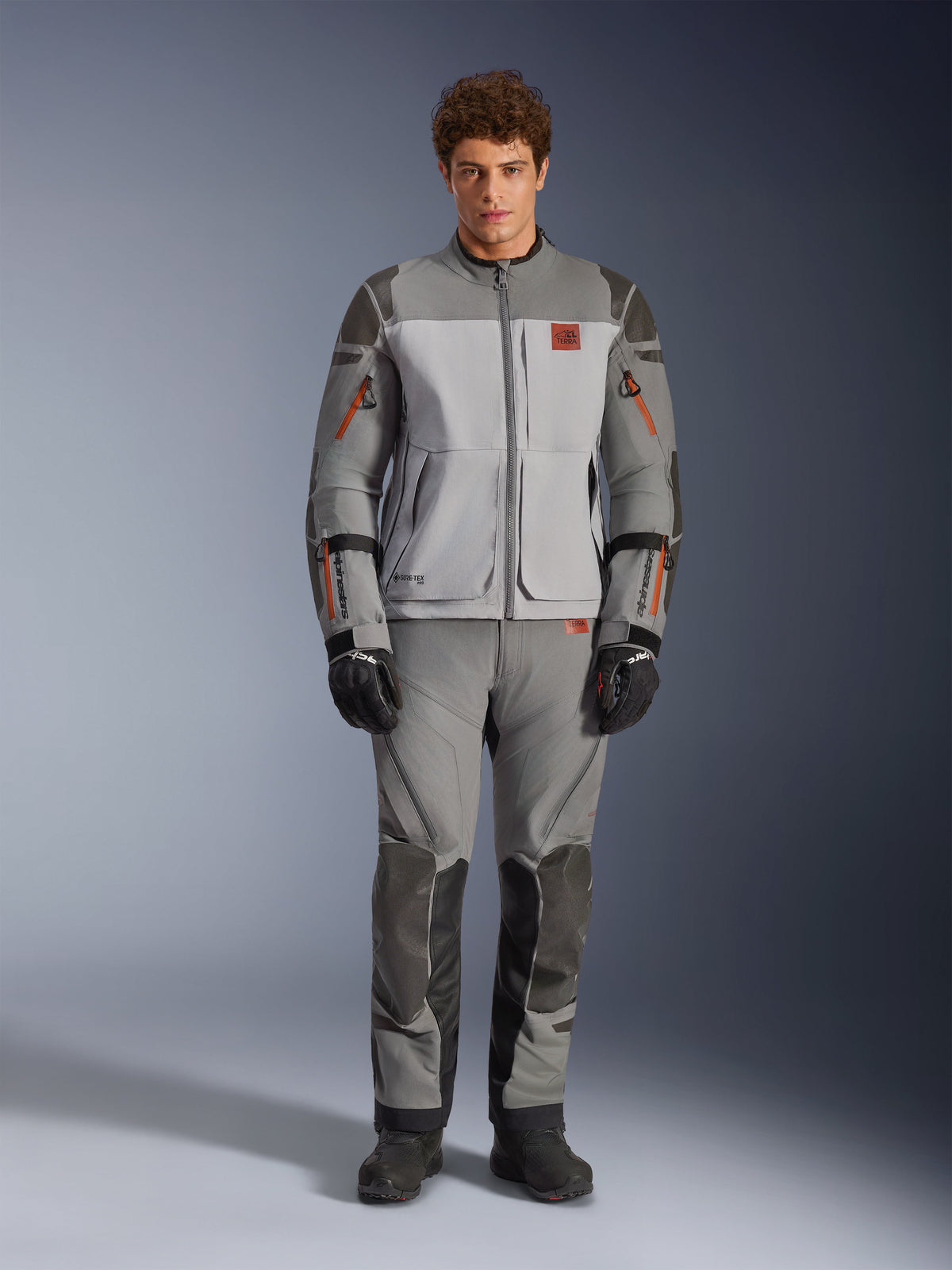 Alpinestars Nazca 3L GORE-TEX PRO Jacket, Adventure Motorcycle Jacket, Dark Gray and Mid Gray, worn by a model with matching pants and black gloves, featuring orange zipper accents and red chest logo, technical protective gear for extreme riding