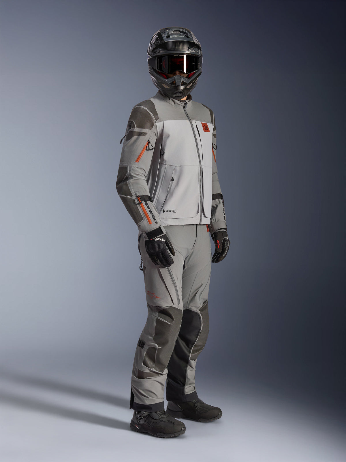 Alpinestars Nazca 3L GORE-TEX PRO Jacket, Adventure Motorcycle Jacket, Dark Gray and Mid Gray, worn as a complete outfit with matching adventure pants, featuring orange accents and Superfabric reinforcements, includes black adventure helmet and gloves, technical apparel for touring and off-road riding