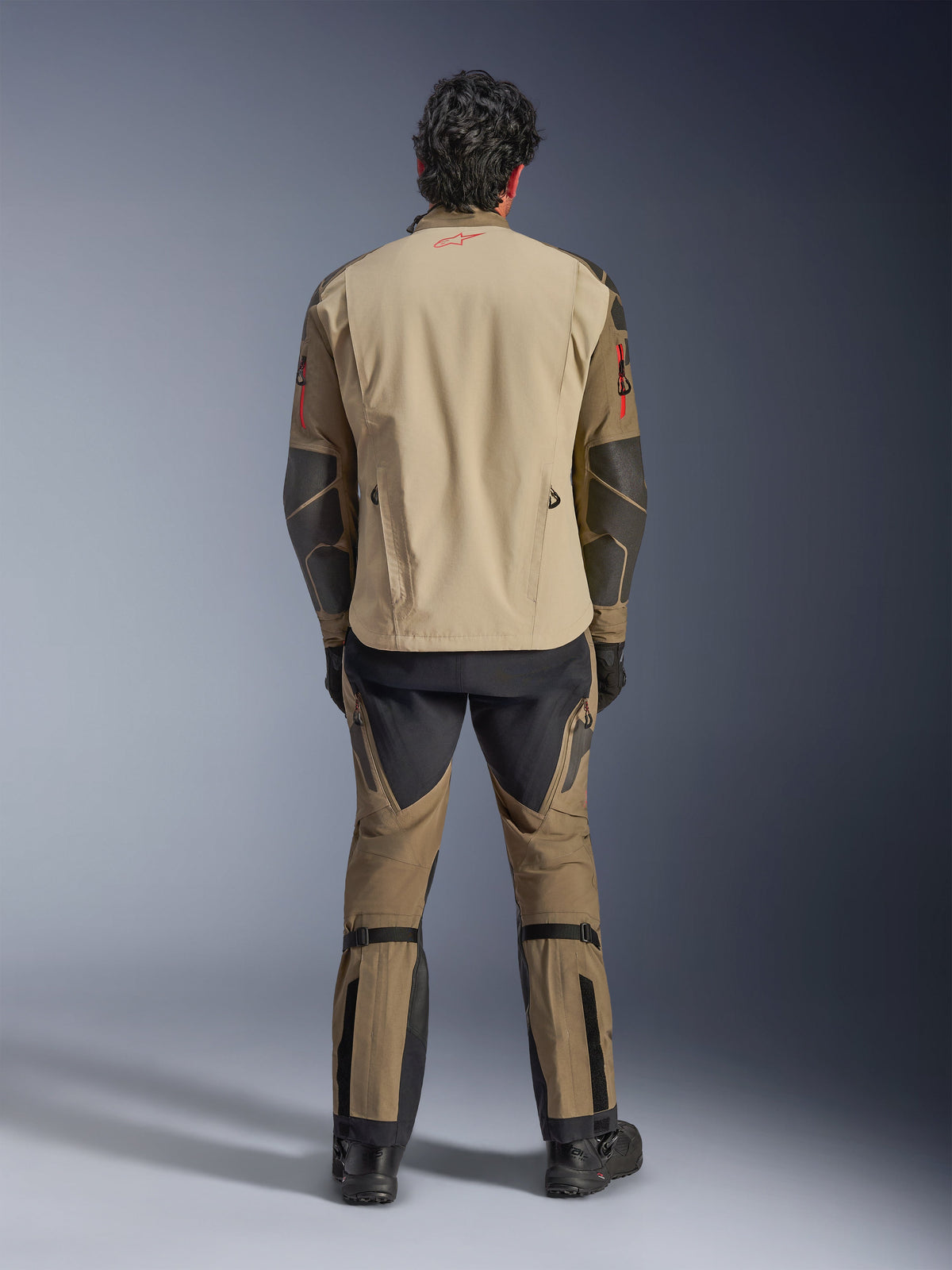 Alpinestars Nazca 3L GORE-TEX PRO Jas in Walnut Rock Brown, achteraanzicht van de rijder, met Superfabric-versterkingen op de ellebogen en ventilatieritsen aan de achterzijde, gedragen met bijpassende adventure-broek, zwarte laarzen en handschoenen, robuuste technische motorkleding voor extreem avontuur.
