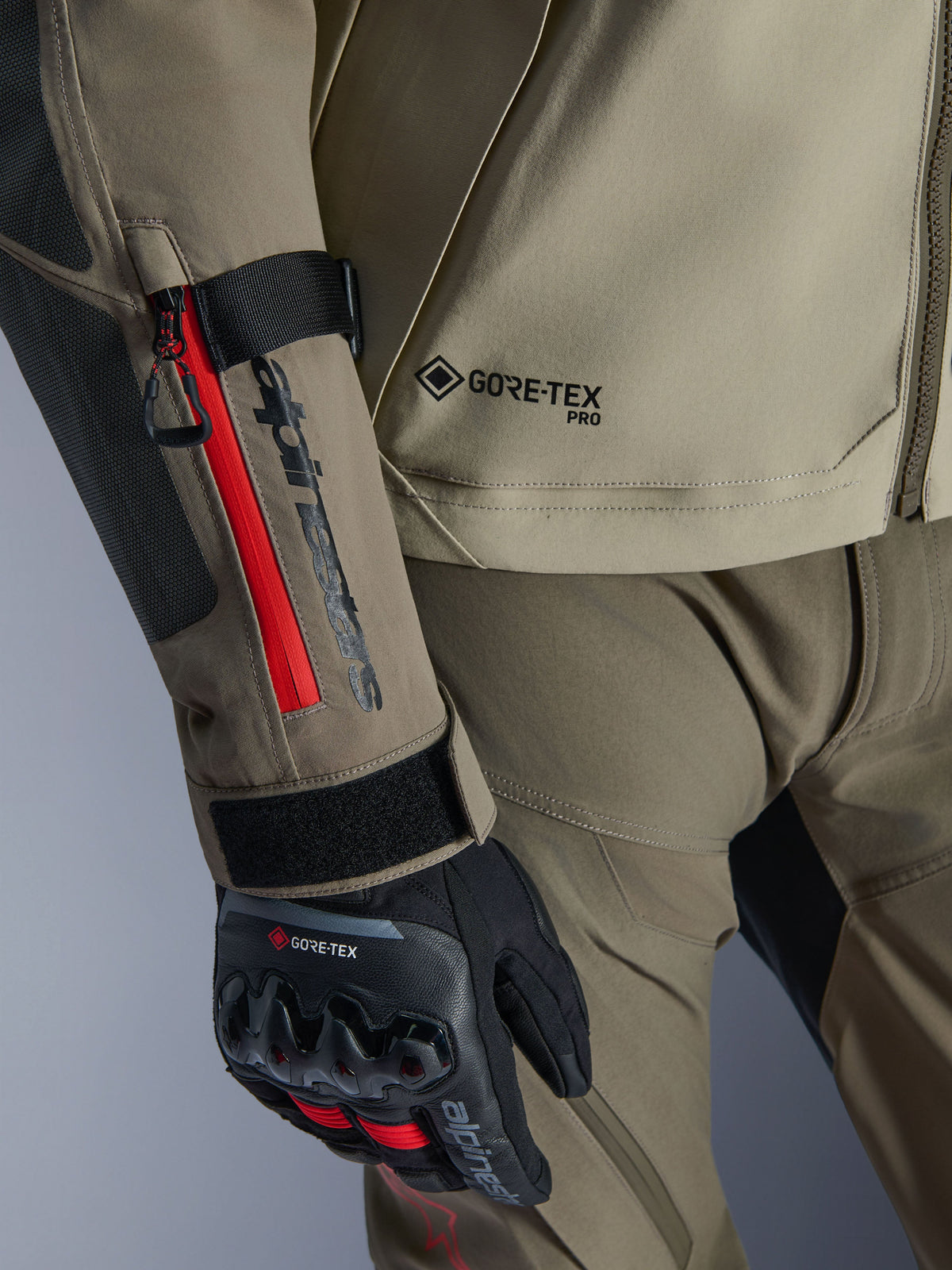Close-up van de Alpinestars Nazca 3L GORE-TEX PRO jas in Walnut Rock Brown, met rode ritsaccenten op de mouw, GORE-TEX PRO-branding en gecombineerd met zwarte GORE-TEX adventure motorhandschoenen.
