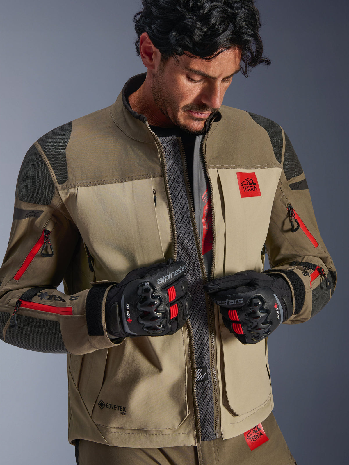 Alpinestars Nazca 3L GORE-TEX PRO Jas, Walnut Rock Brown, adventure motorjas met geopende voorzijde die het centrale mesh-luchtstroompaneel onthult, rode All Terra-branding, gedragen met zwarte GORE-TEX motorhandschoenen, technische uitrusting voor off-road touring