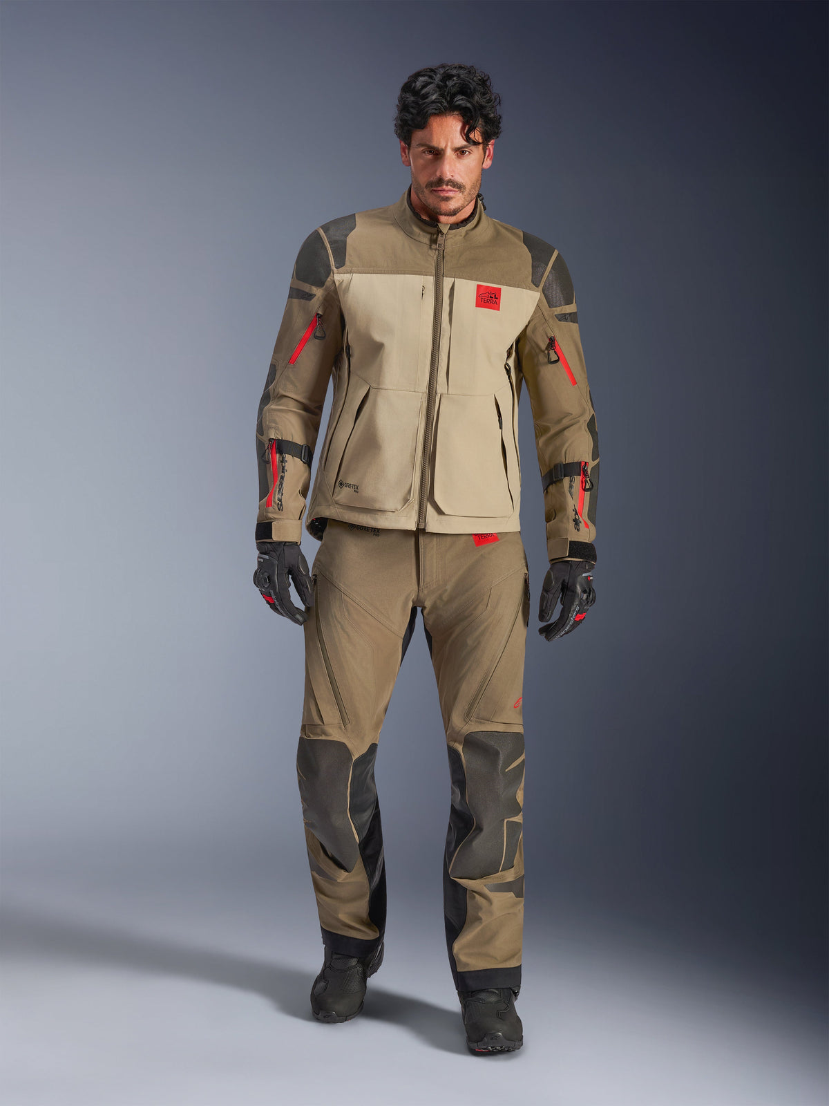Alpinestars Nazca 3L GORE-TEX PRO Jas, Walnut Rock Brown, avontuurlijke motorjas gedragen met bijpassende broek en handschoenen, voorzien van rode ritsaccenten, 'All Terra' logo op de borst en een robuuste textielconstructie.