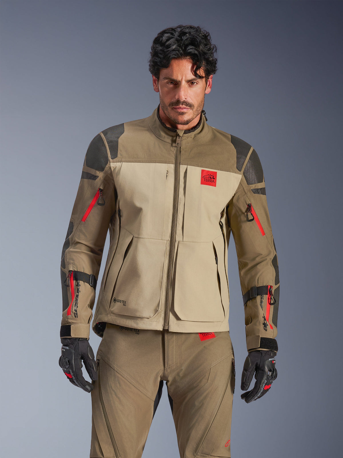 Alpinestars Nazca 3L GORE-TEX PRO jas, adventure motor touringjas, walnootbruin en beige met rode accenten, voorzien van zwarte Superfabric-schouderversterkingen en rode ritsen op de mouwen, gedragen door een rijder met bijpassende uitrusting, premium technische adventure kleding