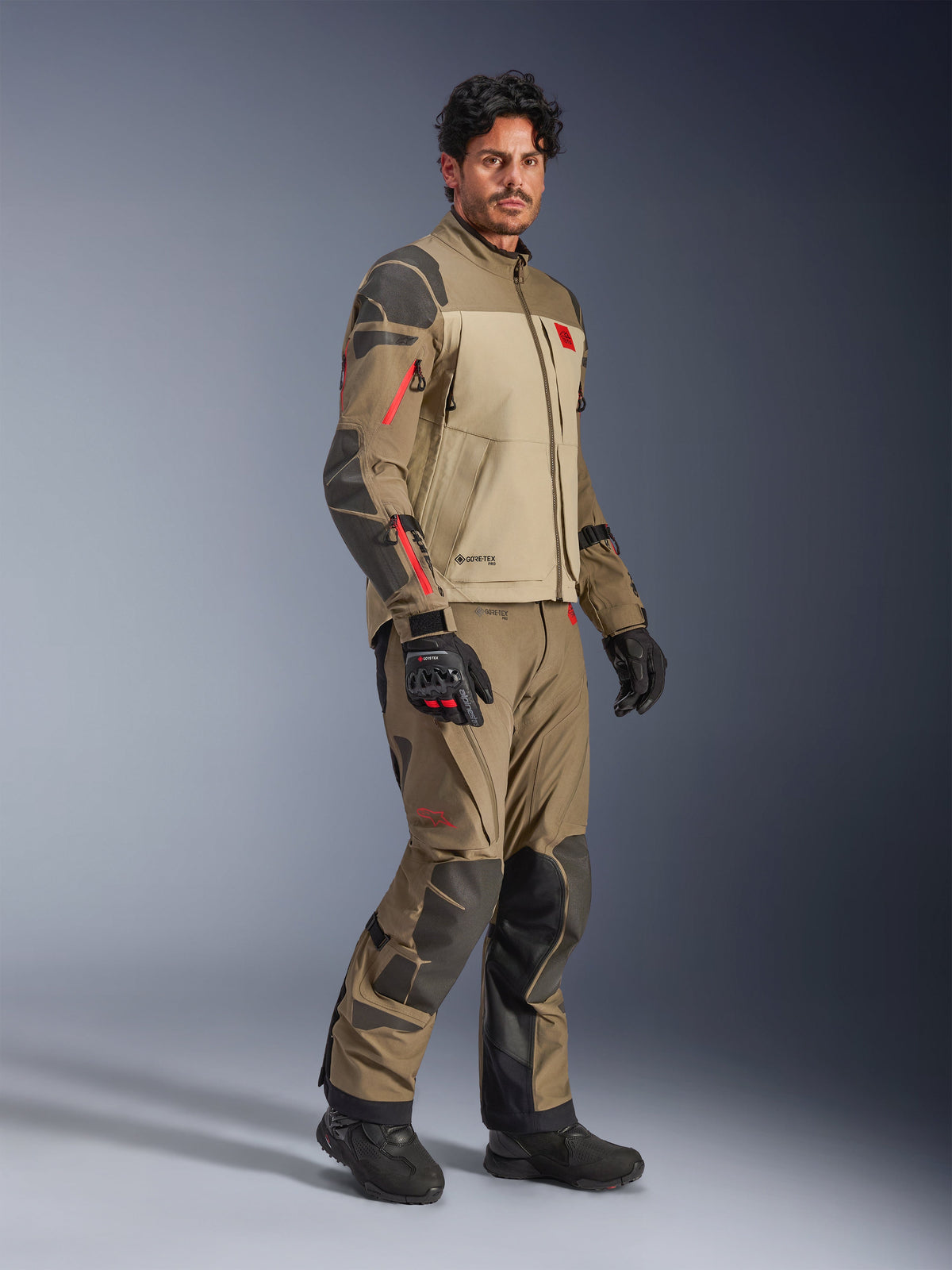 Alpinestars Nazca 3L GORE-TEX PRO jas, Walnut Rock Brown, adventure motorjas gedragen met bijpassende broek en zwarte handschoenen, voorzien van donkergrijze verstevigingen, rode ritsdetails en een hoogwaardige gelamineerde buitenlaag.