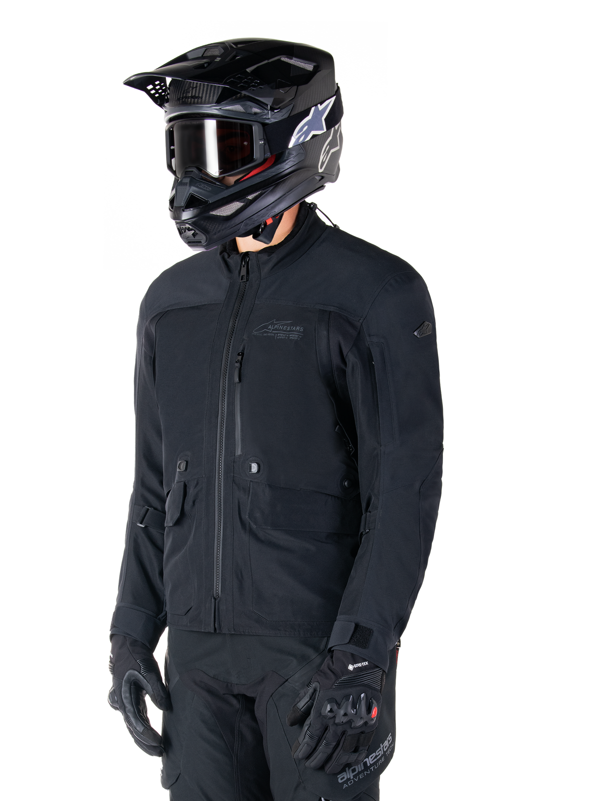Alpinestars St-7 Prime GORE-TEX jas, zwarte motorjas, gedragen door een rijder met zwarte helm en motorbril, voorzien van grote cargozakken op de borst en een gelamineerde waterdichte buitenlaag, premium uitrusting voor adventure touring en woon-werkverkeer