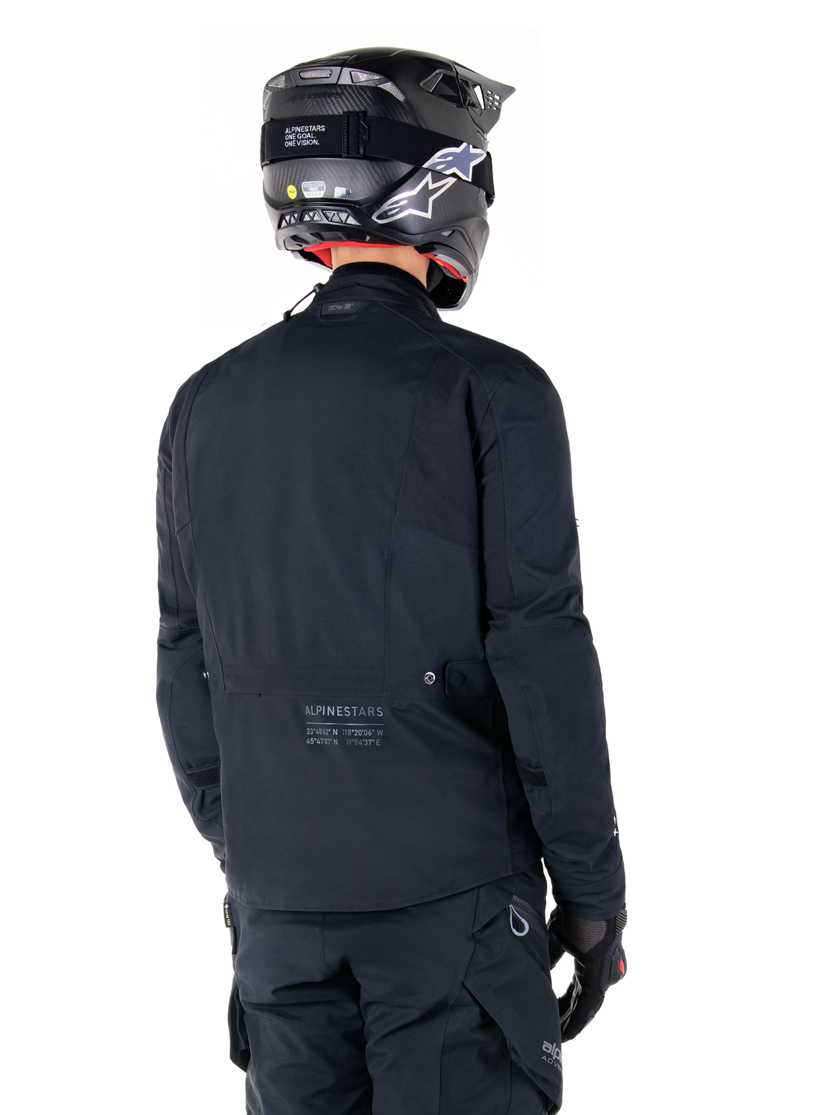 Alpinestars St-7 Prime GORE-TEX Jas, zwart, achteraanzicht van een rijder die de jas draagt met een bijpassende helm en broek, voorzien van Alpinestars-branding en geografische coördinaten op de onderrug.