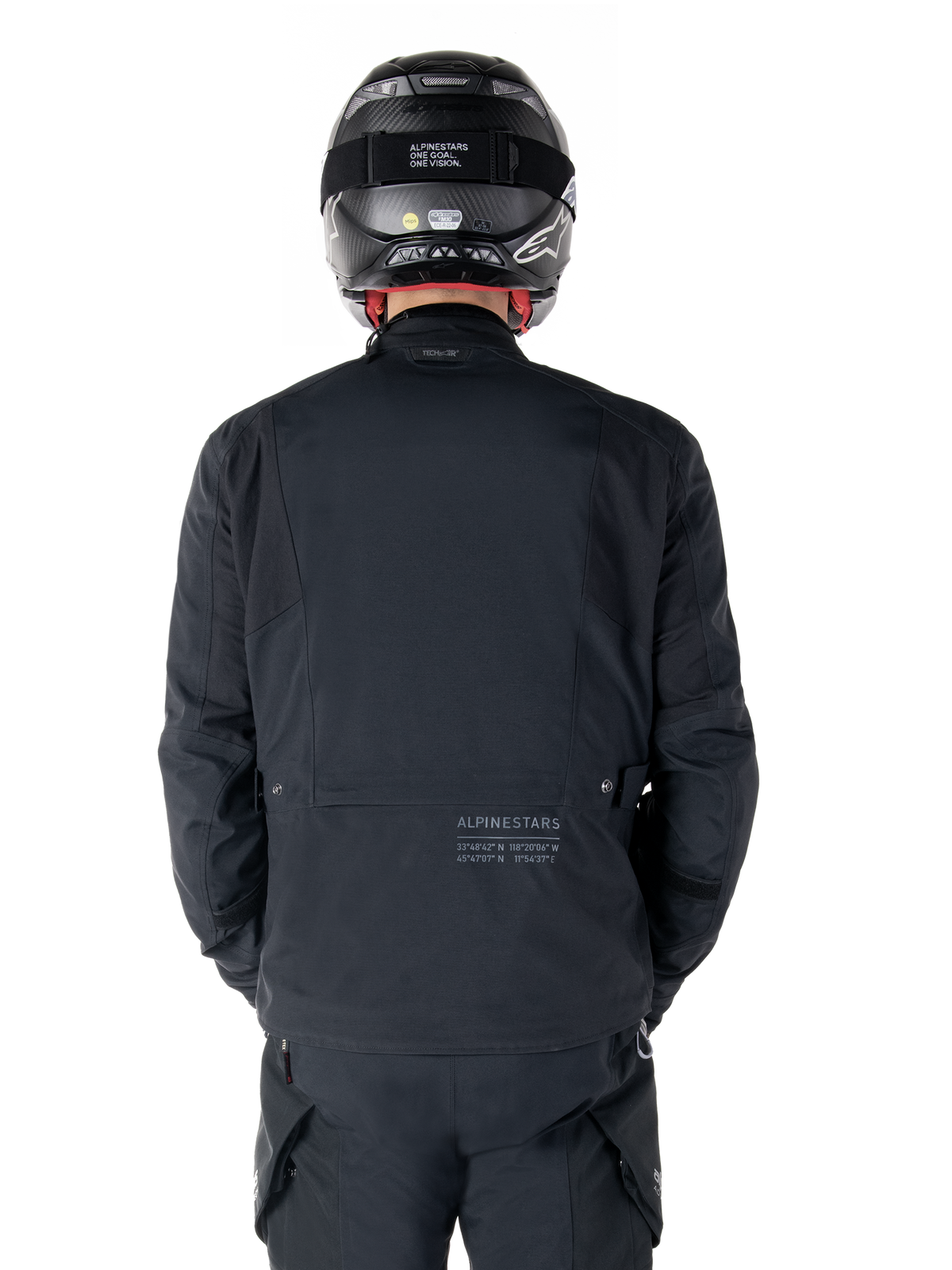 Alpinestars St-7 Prime GORE-TEX Jas, zwart, achteraanzicht van een motorrijder, met subtiel logo en geografische coördinaten op de onderrug, gedragen met een bijpassende zwarte helm en broek, strakke en technische motorkleding