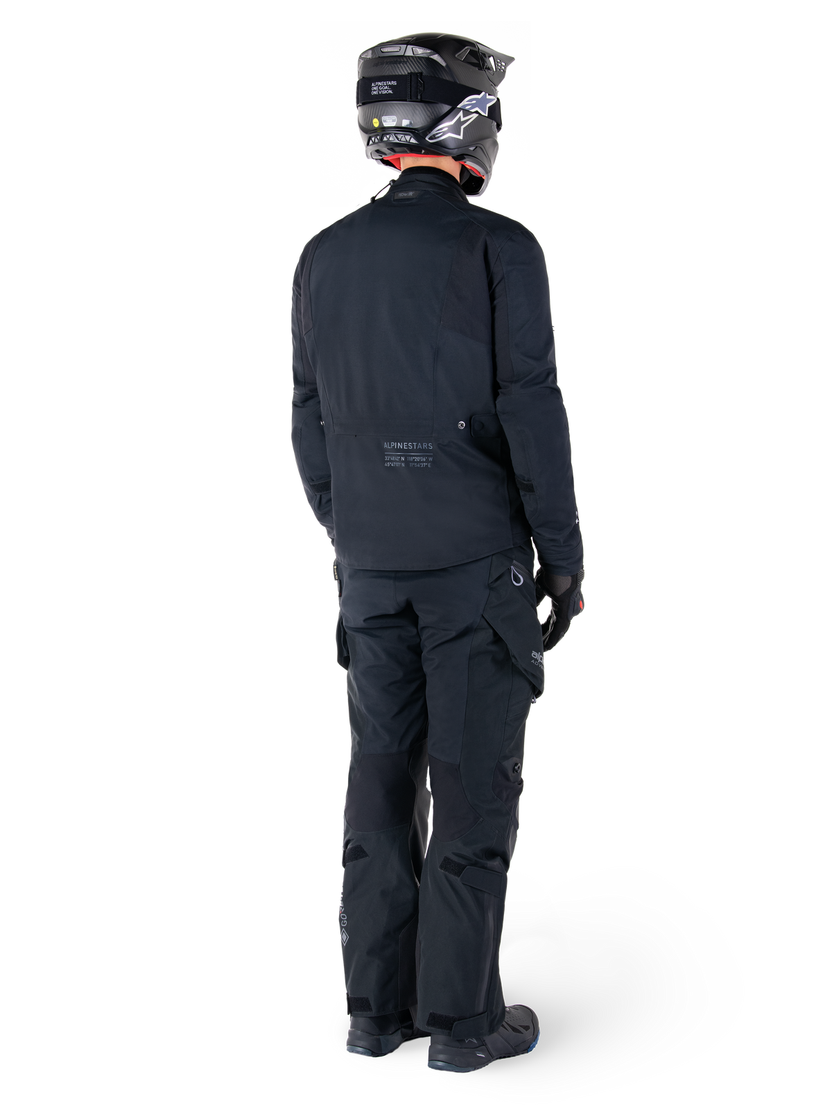 Achteraanzicht van een motorrijder die de Alpinestars St-7 Prime GORE-TEX Jas in het zwart draagt, gecombineerd met een bijpassende zwarte toerbroek, een zwarte motorhelm met wit logo en zwarte motorlaarzen, volledige beschermende adventure-uitrusting voor toeren en woon-werkverkeer.