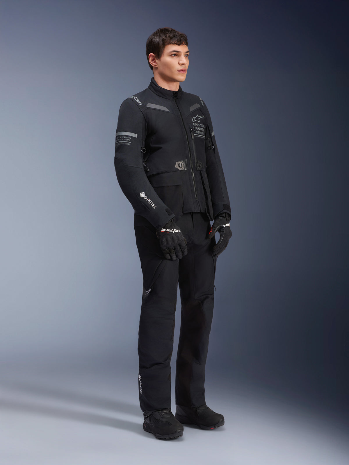 Alpinestars ST-7 2L GORE-TEX broek, gedragen door een model met de bijbehorende ST-7 jas, volledig zwarte adventure touring motoroutfit, voorzien van GORE-TEX branding, cargozakken, zwarte handschoenen en laarzen, studiofoto tegen een grijze achtergrond