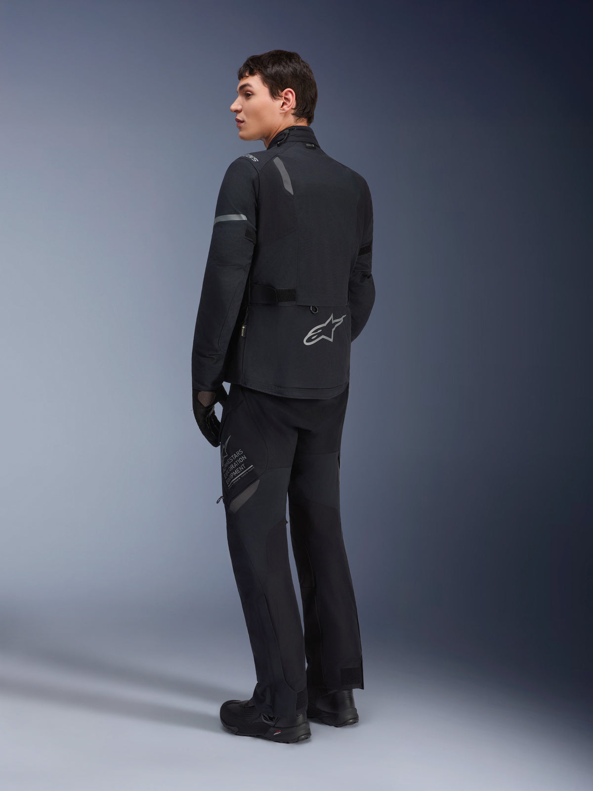 Alpinestars ST-7 2L GORE-TEX broek, adventure touring broek, zwart en donkergrijs, gedragen met bijpassende ST-7 jas en zwarte laarzen, voorzien van 'Alpinestars Exploration Equipment' branding op de dij, ontworpen voor waterdicht en ademend rijden in vier seizoenen