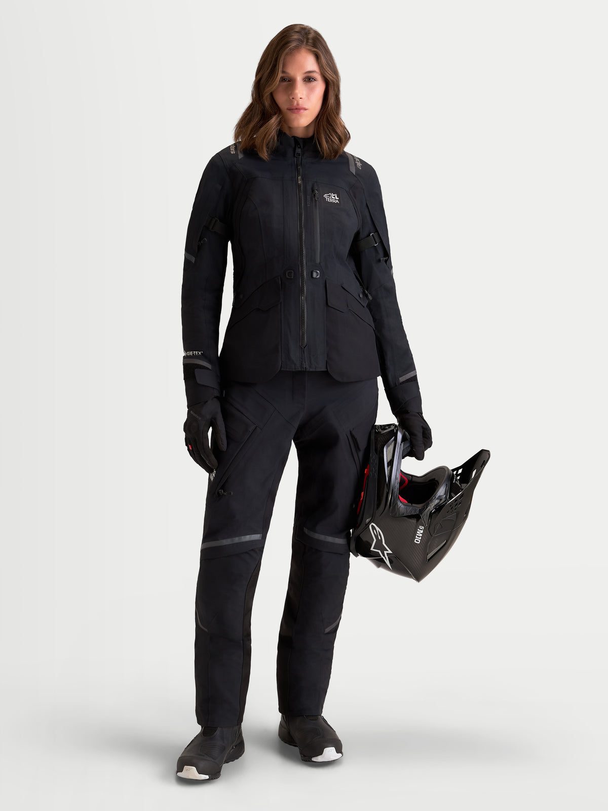 Alpinestars Stella ST-7 V2 2L GORE-TEX jas, adventure motorjas voor dames in het zwart, volledig overzicht op een model dat bijpassende kleding draagt en een helm vasthoudt, waterdichte textielconstructie met CE-gecertificeerde bescherming