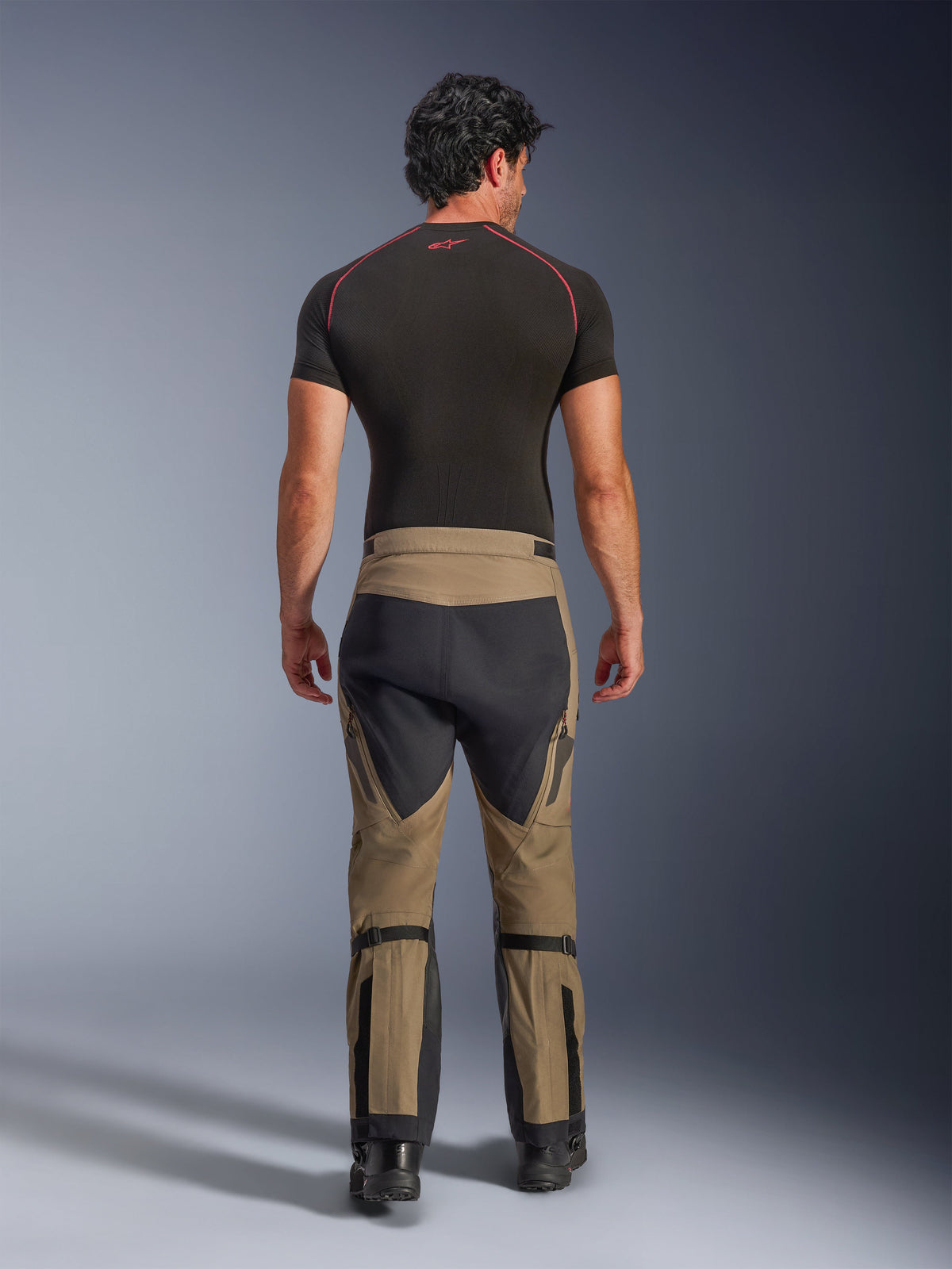 Alpinestars Nazca 3L GORE-TEX PRO Broek, Walnut Black adventure motorbroek, achteraanzicht op een model met een zwarte basislaag en laarzen, voorzien van een tweekleurig ontwerp met kuitverstellers en stretchpanelen.