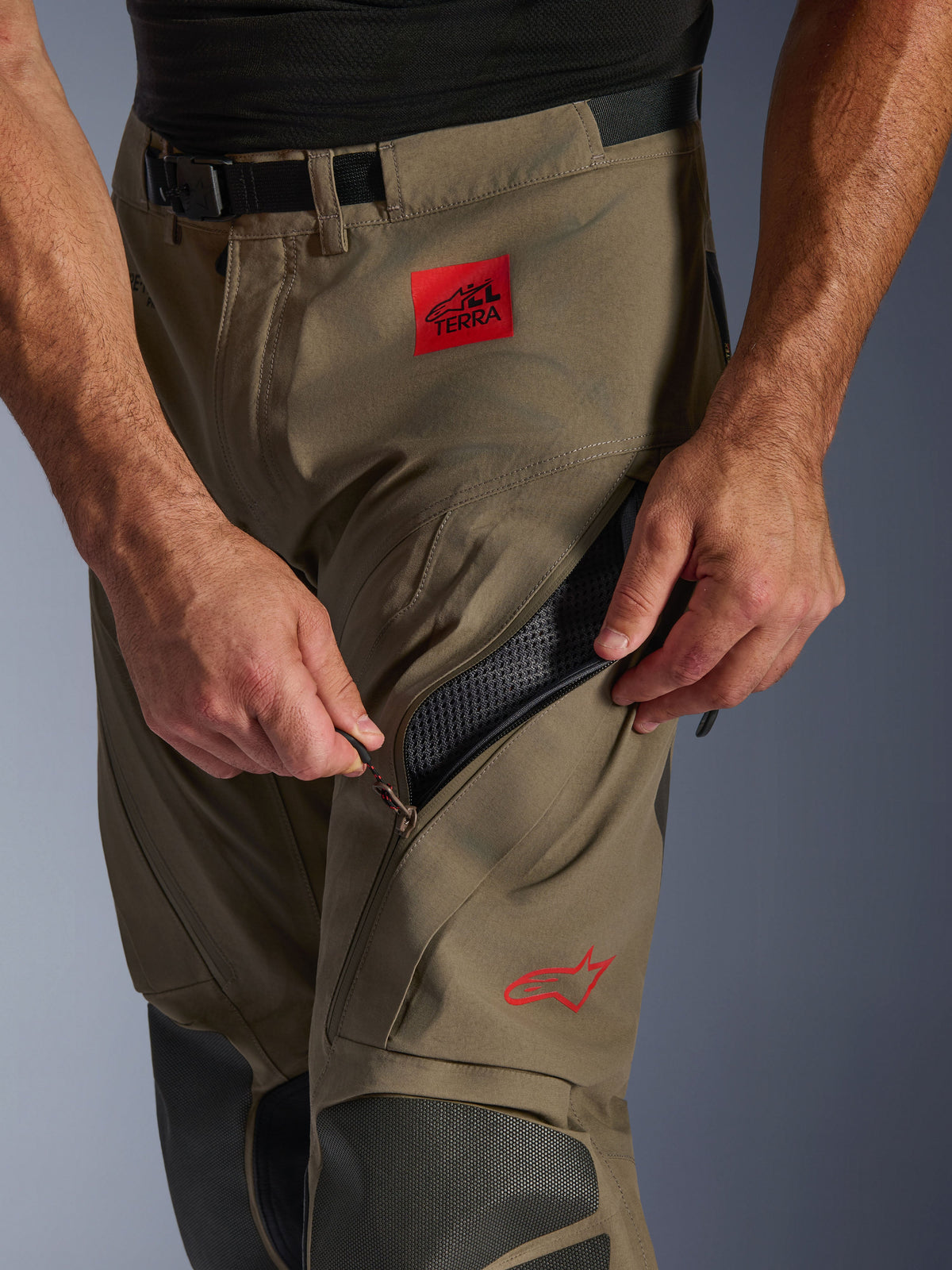 Alpinestars Nazca 3L GORE-TEX PRO broek in Walnut Black, close-up detail van het dijgedeelte waarbij een hand de grote ventilatieopening met ritssluiting opent, voorzien van een rode ALL TERRA-patch en rood Alpinestars-logo, technische adventure motoruitrusting.